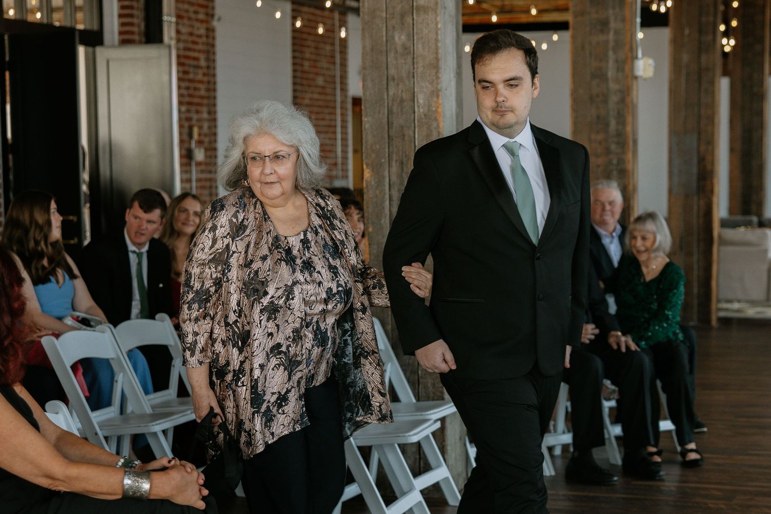 Young man in suit escorting elderly woman down the aisle at a wedding ceremony.