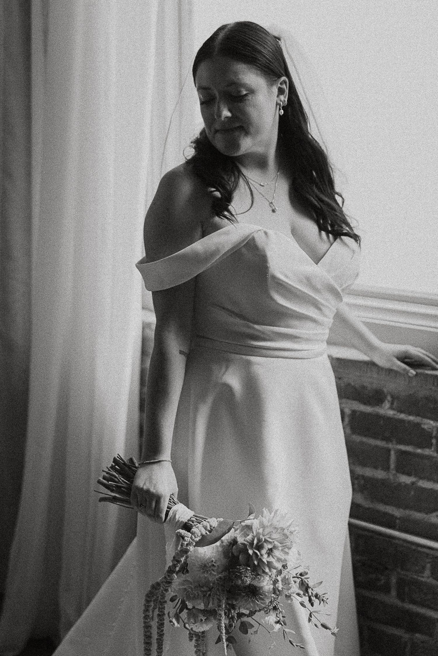Black and white portrait of bride in off-shoulder gown holding bouquet near a curtained window.