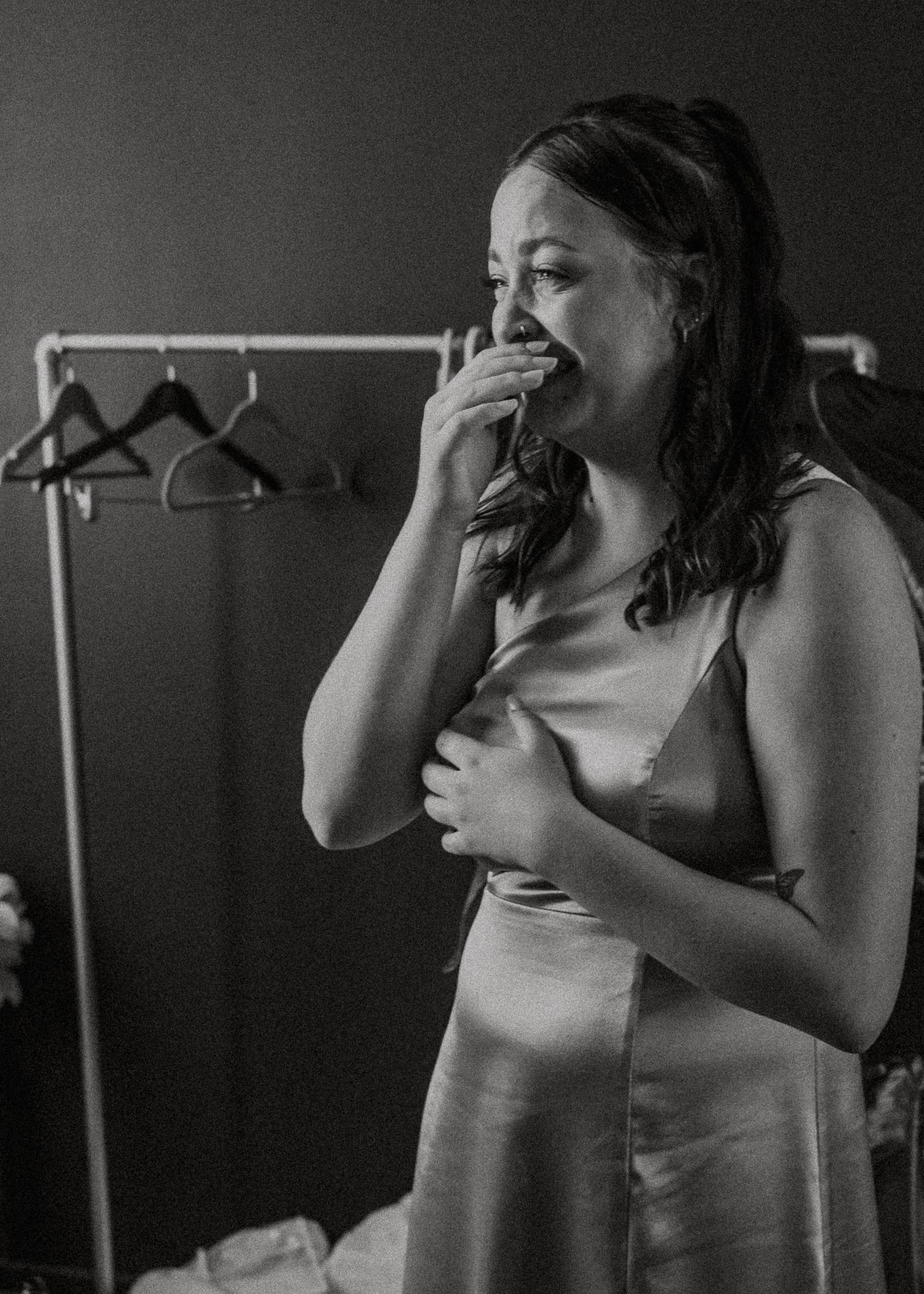 Black and white candid photo of emotional bridesmaid in satin slip dress near a clothing rack.