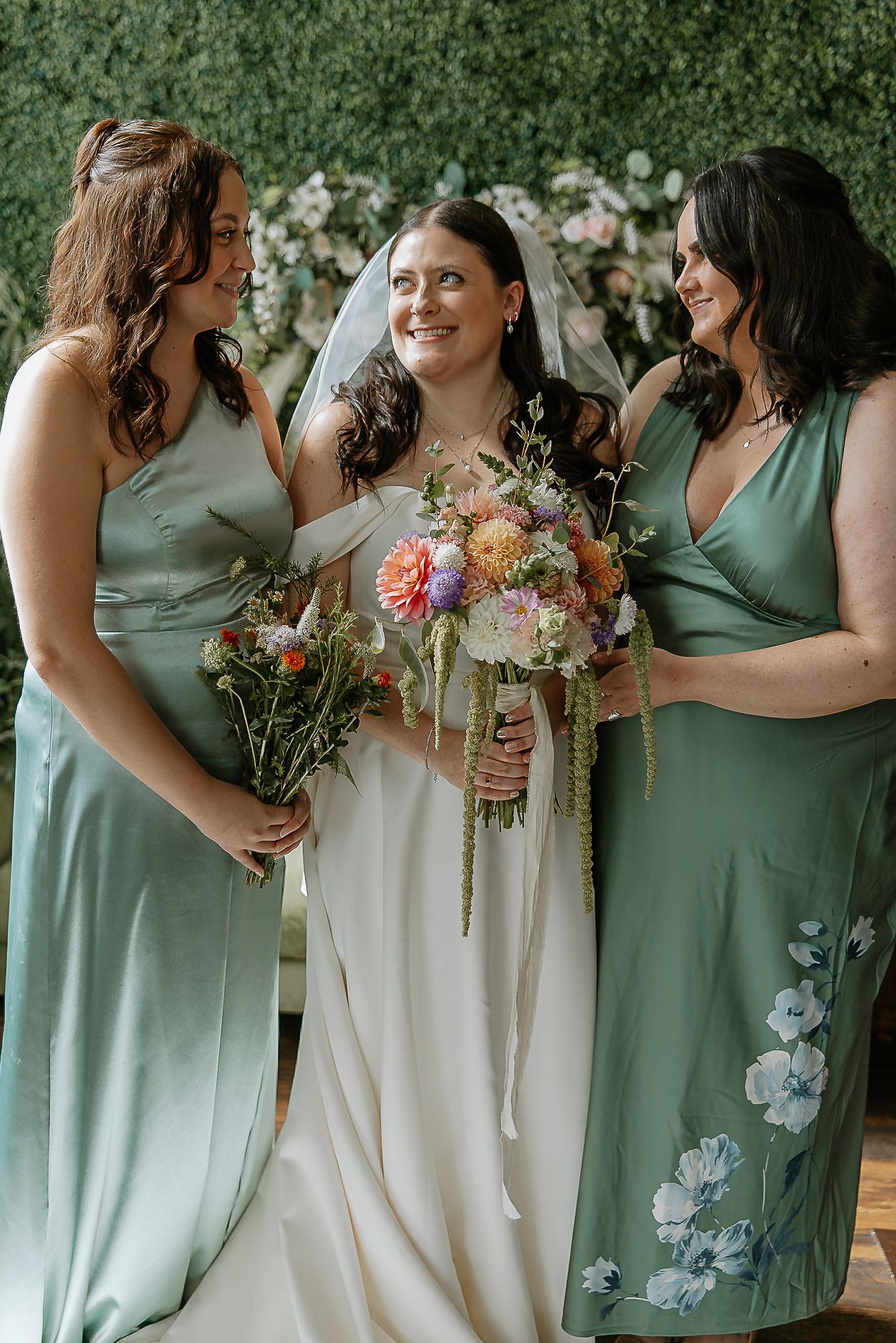 Bride with veil and colorful bouquet laughs with two bridesmaids in sage green dresses at wedding.