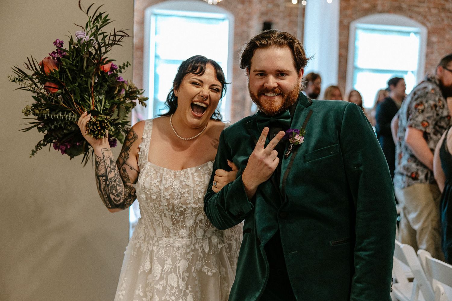 Excited bride and groom celebrate after wedding ceremony, bride showing tattoos and holding bouquet.