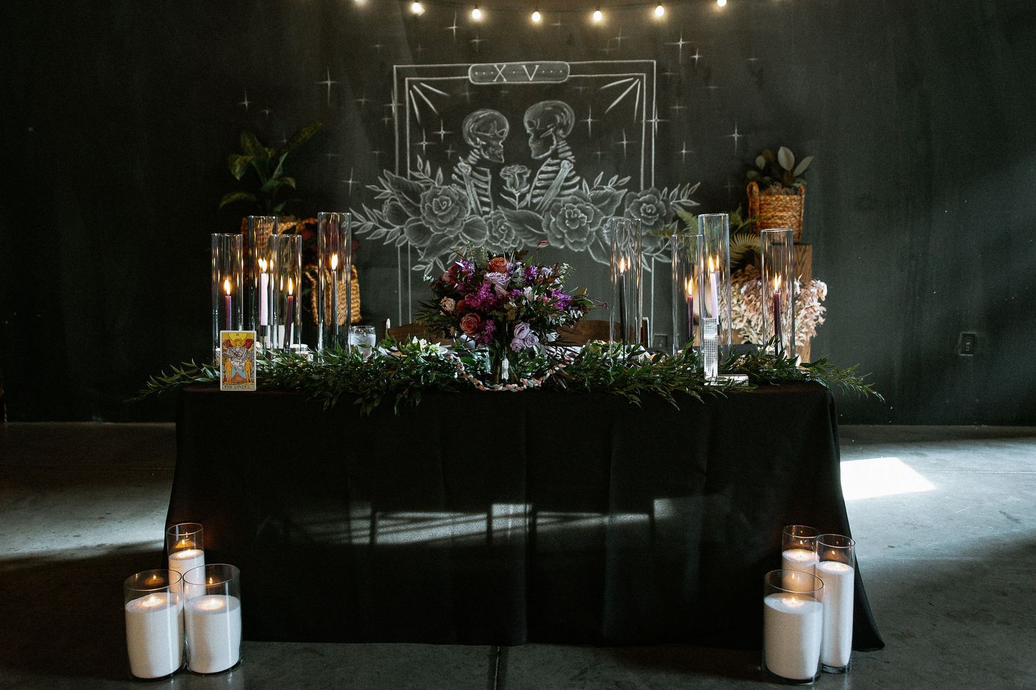 Gothic wedding sweetheart table with black linen, tarot skeleton chalk art backdrop, and floral centerpiece.