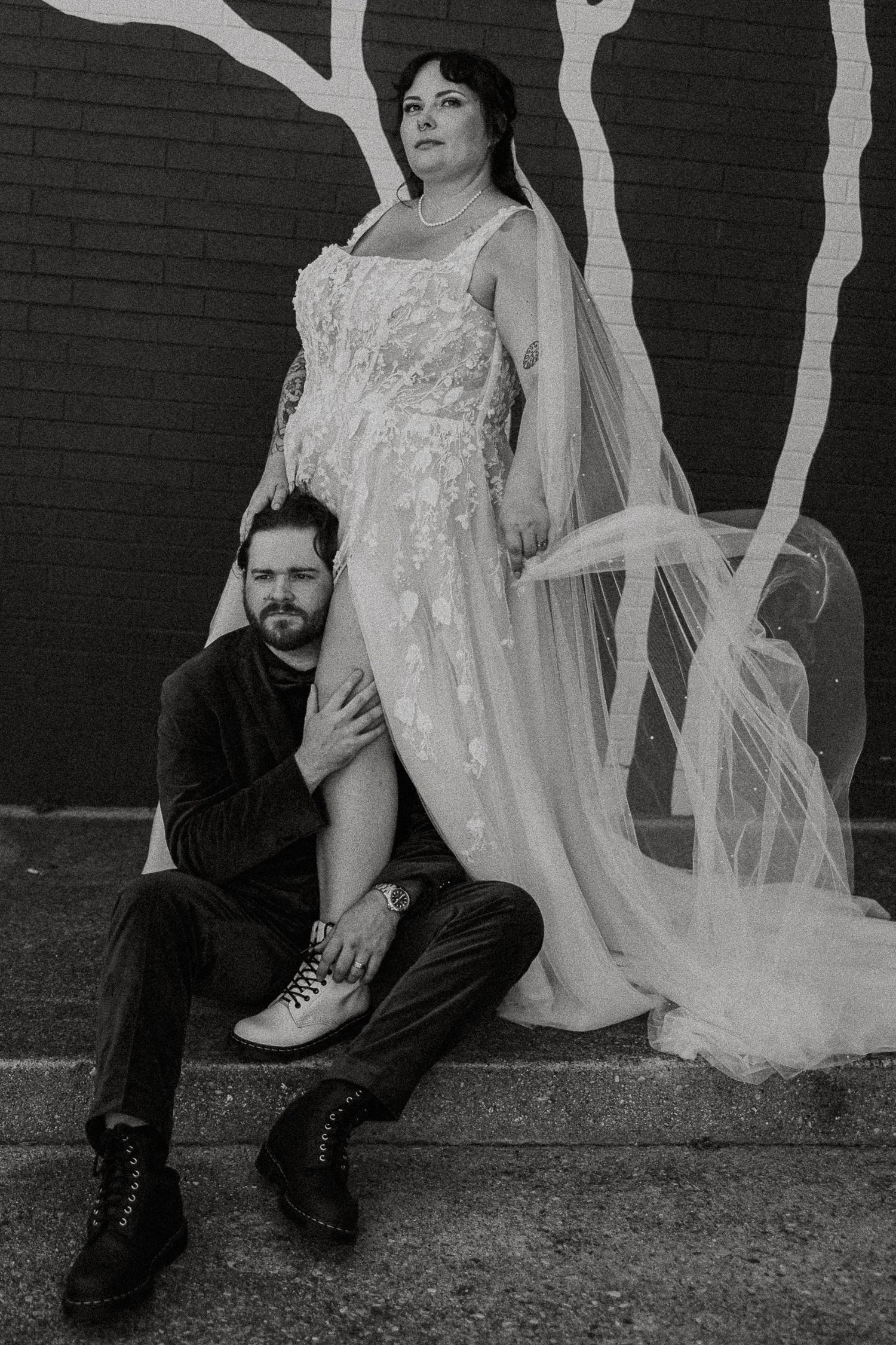 Black and white wedding portrait of bride standing over seated groom against graffiti brick wall.