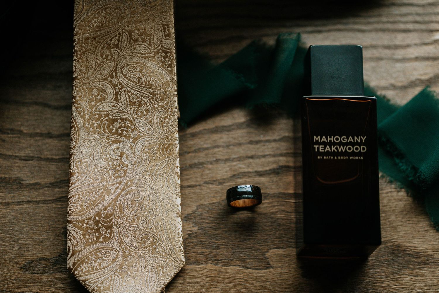 Gold paisley tie, Mahogany Teakwood cologne, green bow tie, and a black ring arranged on a rustic wooden surface.