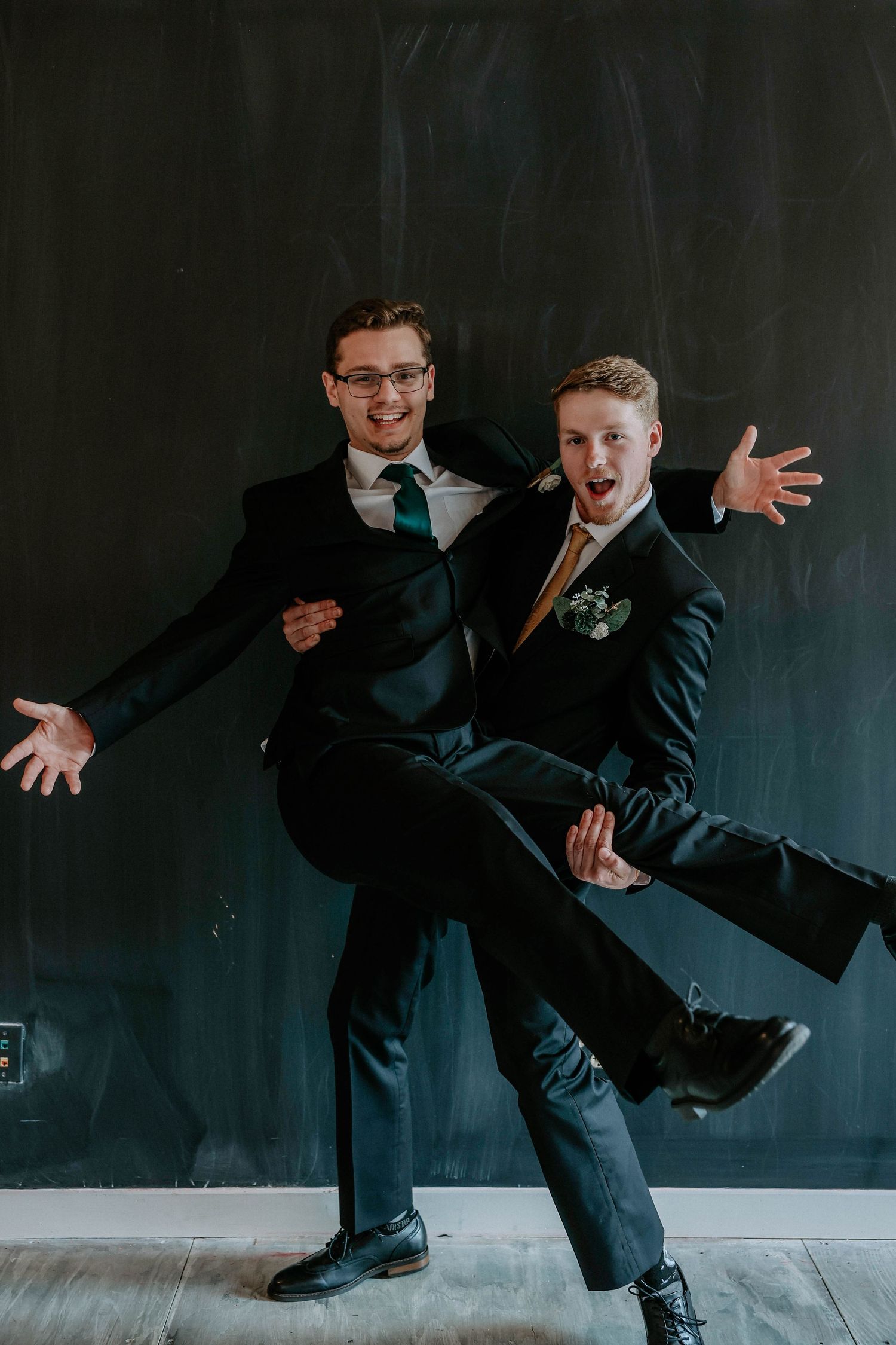 Two young men in dark suits strike a fun pose against a chalkboard wall at a formal event.