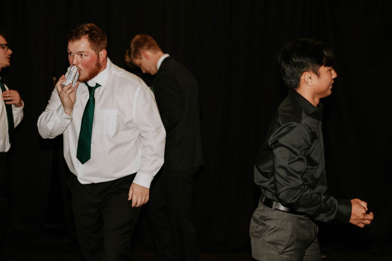 Men in formal attire at an event, one drinking from a glass, wearing white shirts and ties.