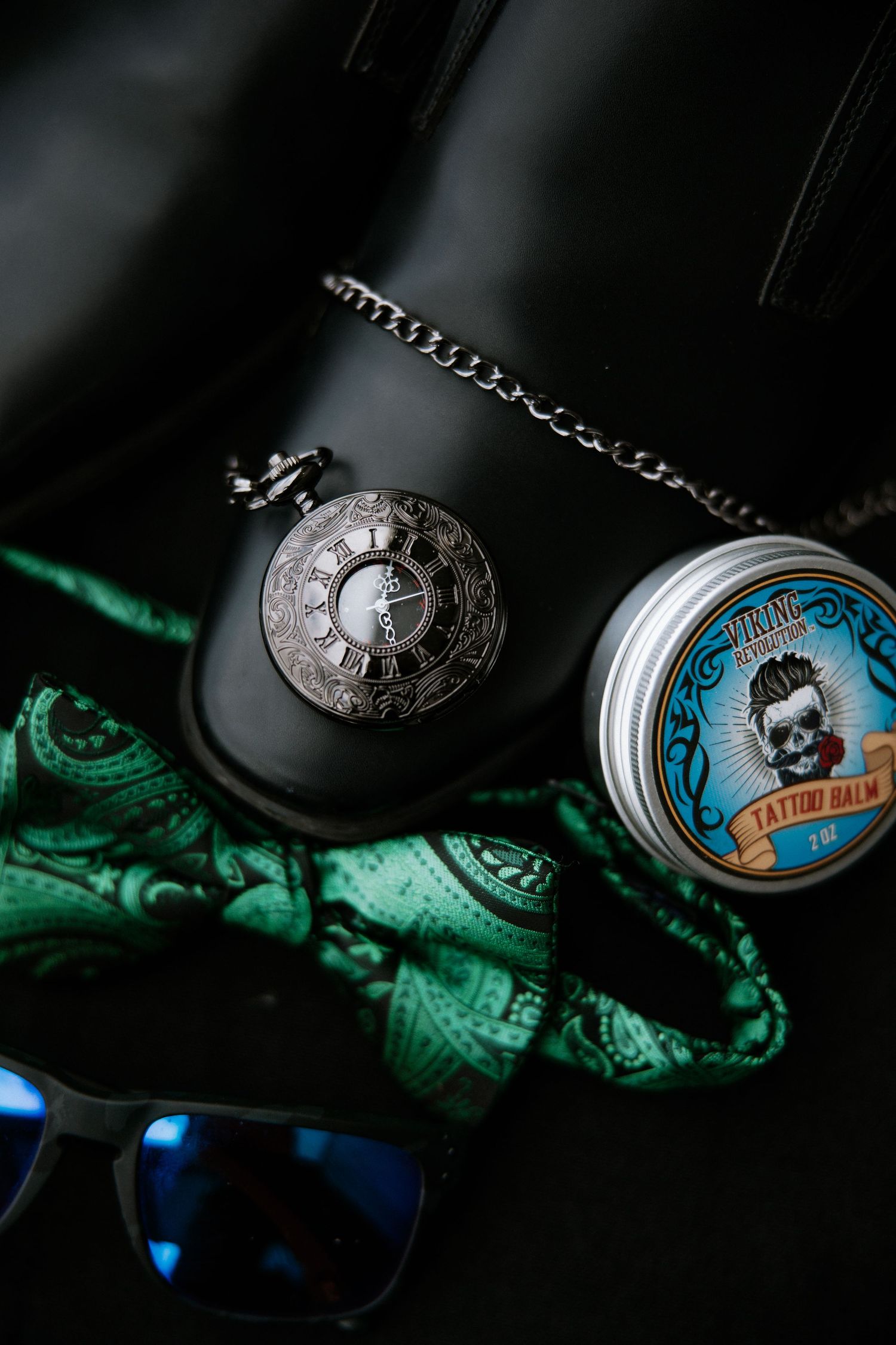 Pocket watch, Viking tattoo balm tin, green bandana, and sunglasses arranged on black leather bag.