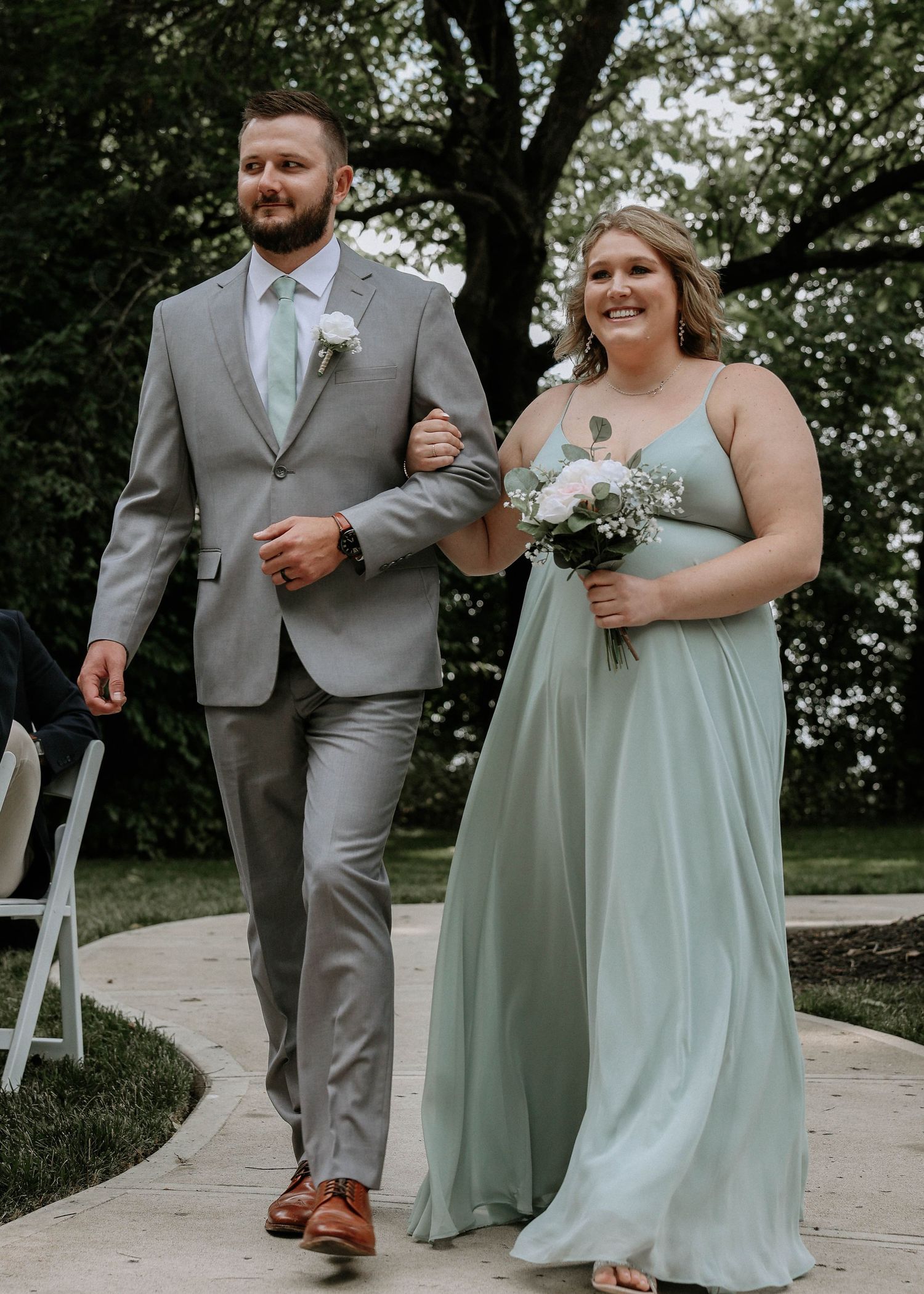 A groomsman in a gray suit escorts a bridesmaid in a sage green gown down the aisle at an outdoor wedding.