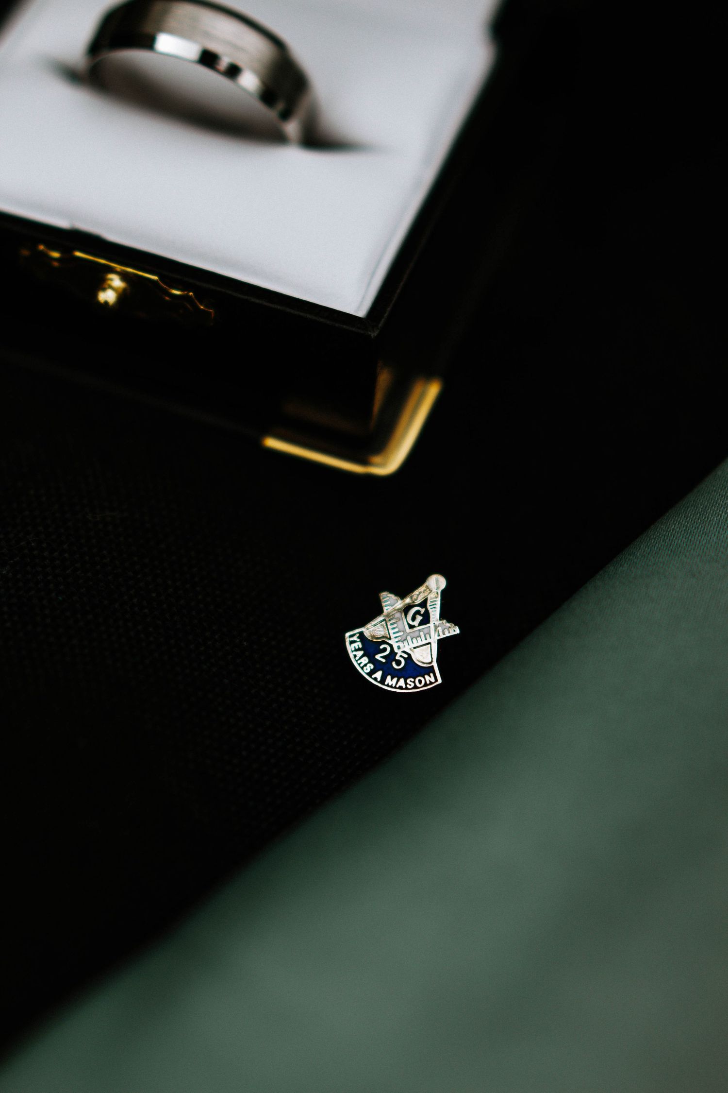 Masonic lodge pin beside a silver wedding band in a white ring box on dark surface.