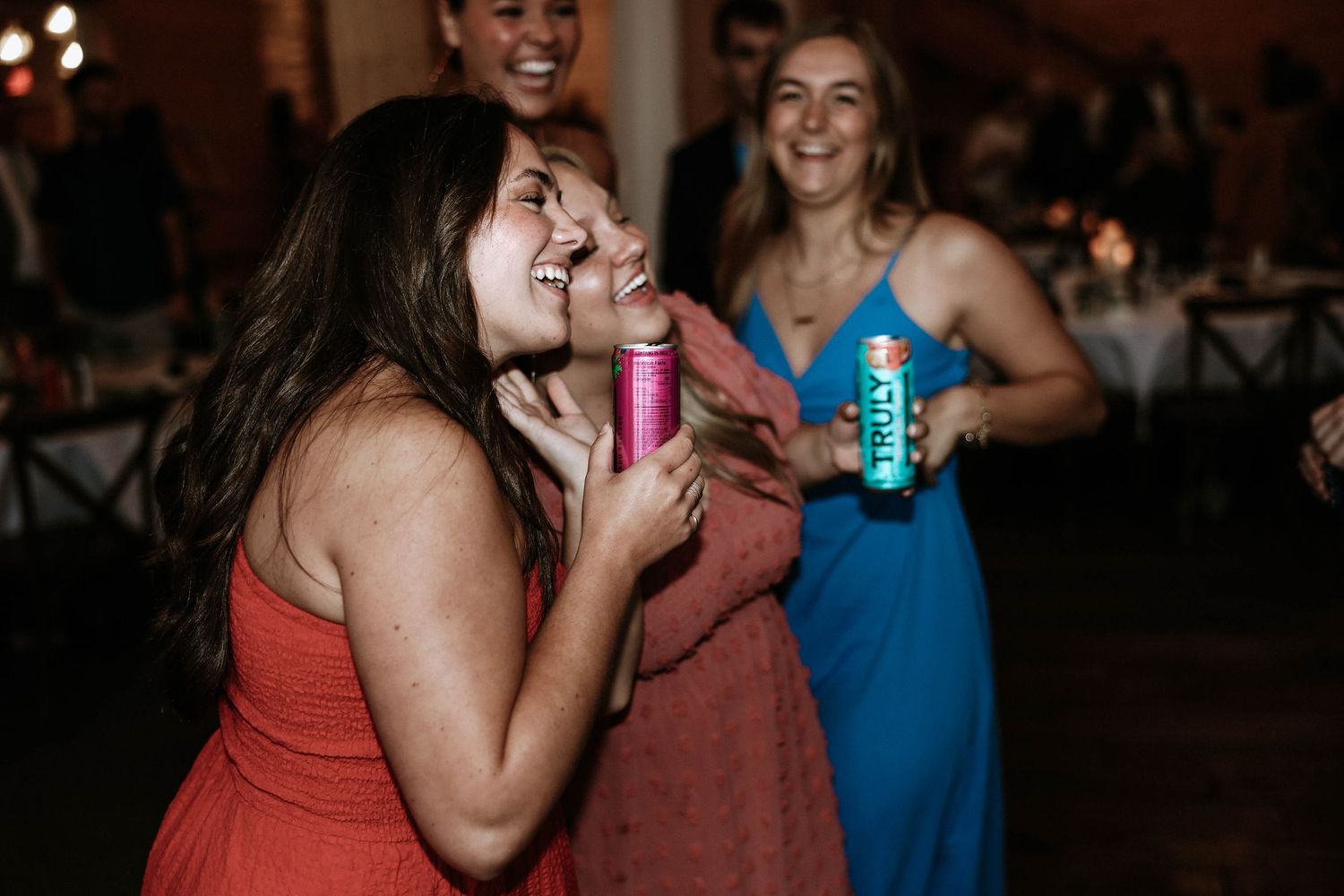 Women laughing and dancing at a wedding reception party, holding colorful canned drinks.