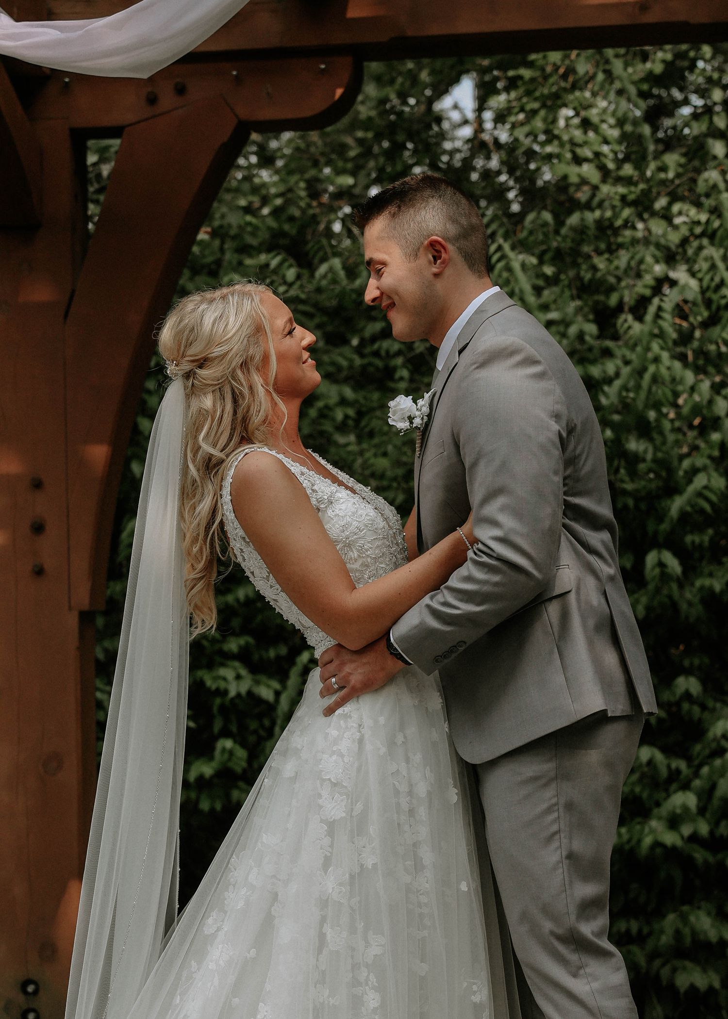 Smiling bride in lace gown and groom in gray suit share a tender moment under a wooden arbor.