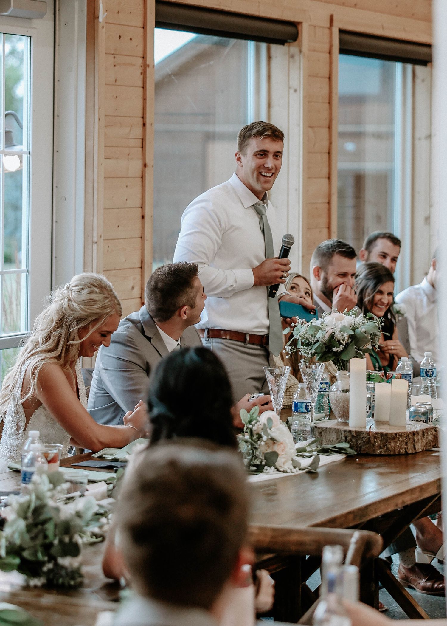 Best man giving a heartfelt wedding speech to laughing guests at rustic barn reception.