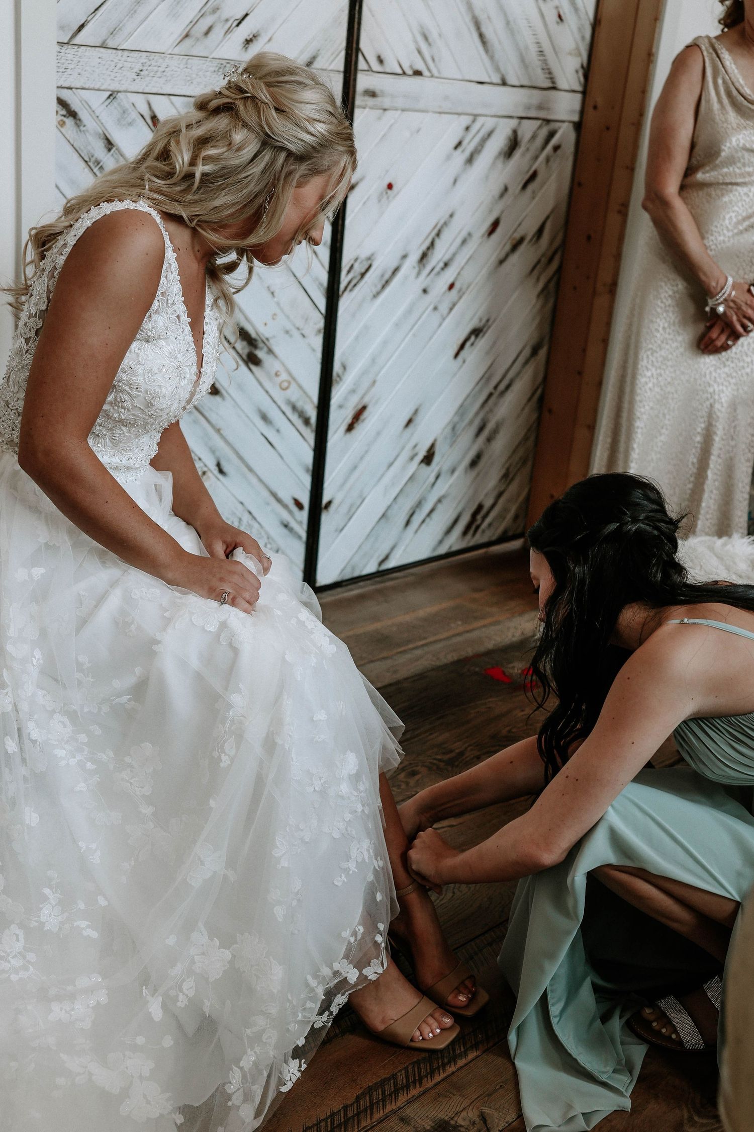 Bridesmaid helping bride with shoes while wearing white lace wedding gown before ceremony.