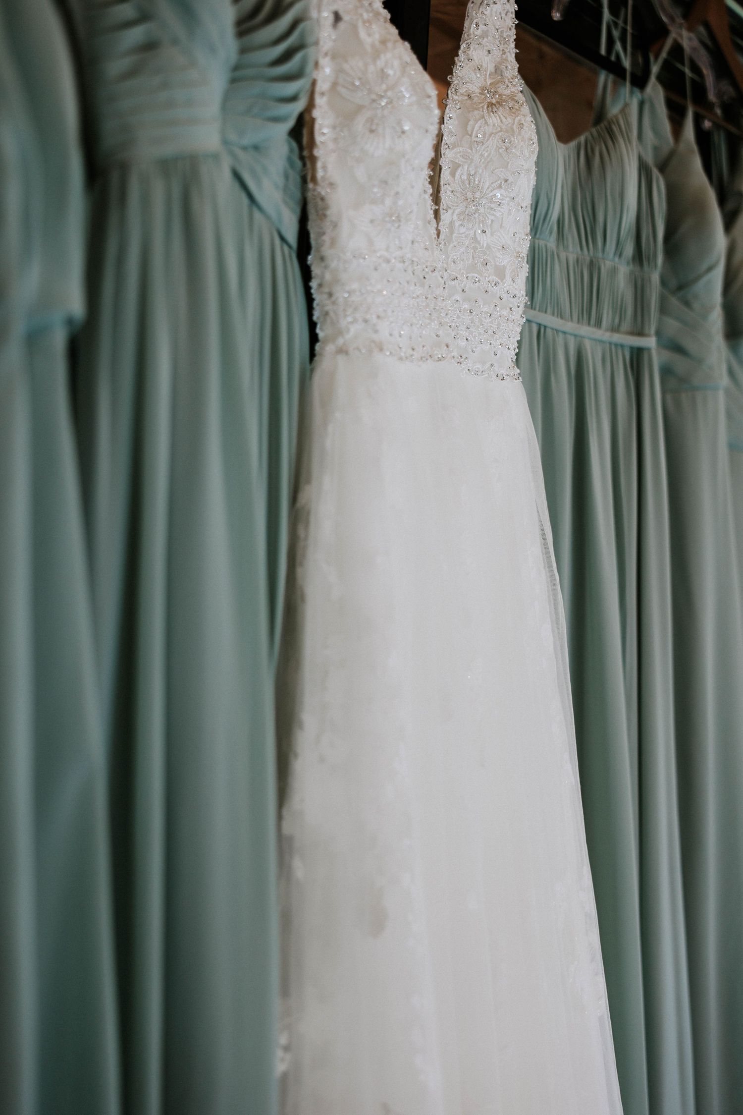 White beaded lace wedding gown hanging beside sage green chiffon bridesmaid dresses on a rack.