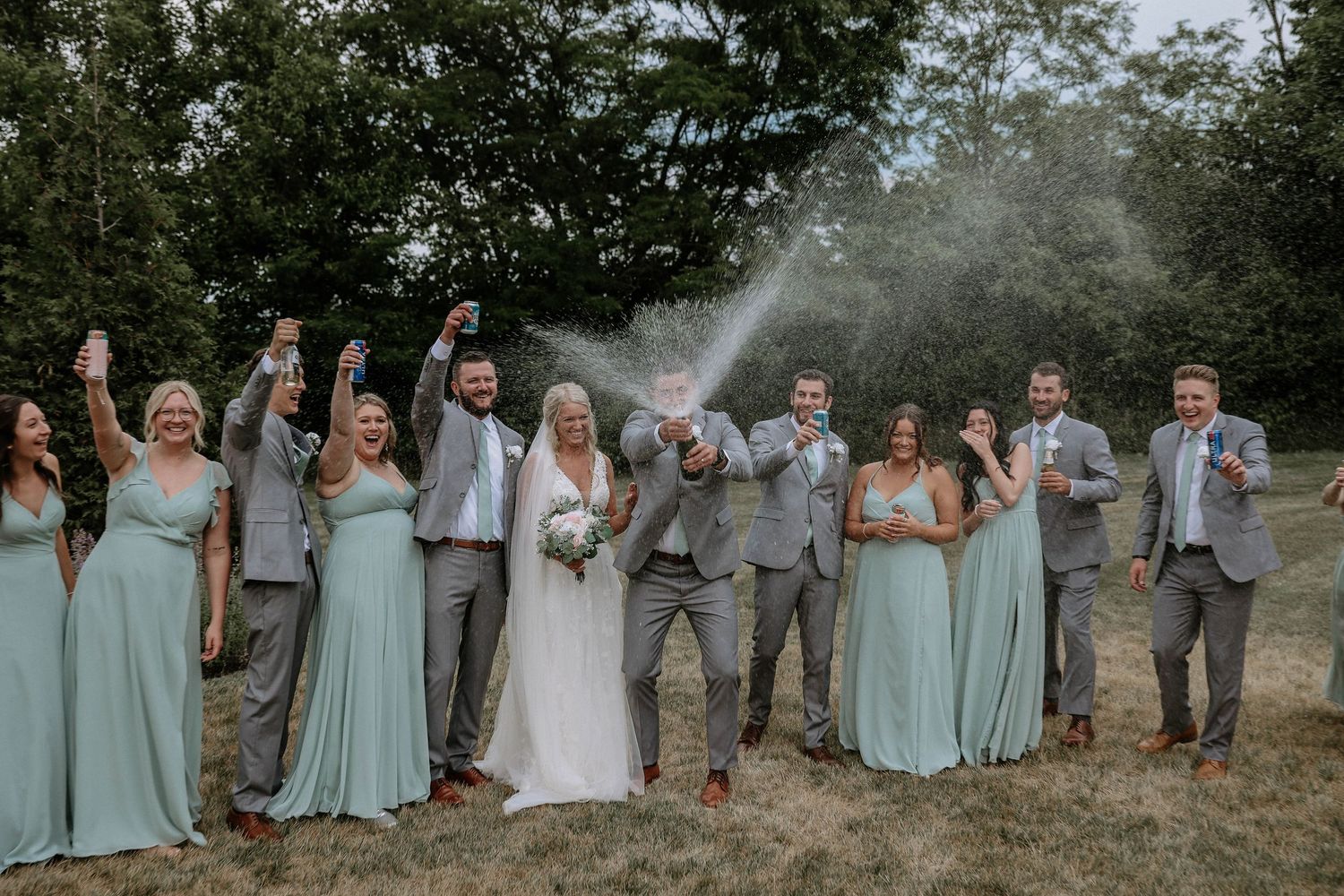Wedding party celebrating with champagne spray outdoors, bridesmaids in sage green gowns and groomsmen in gray suits.