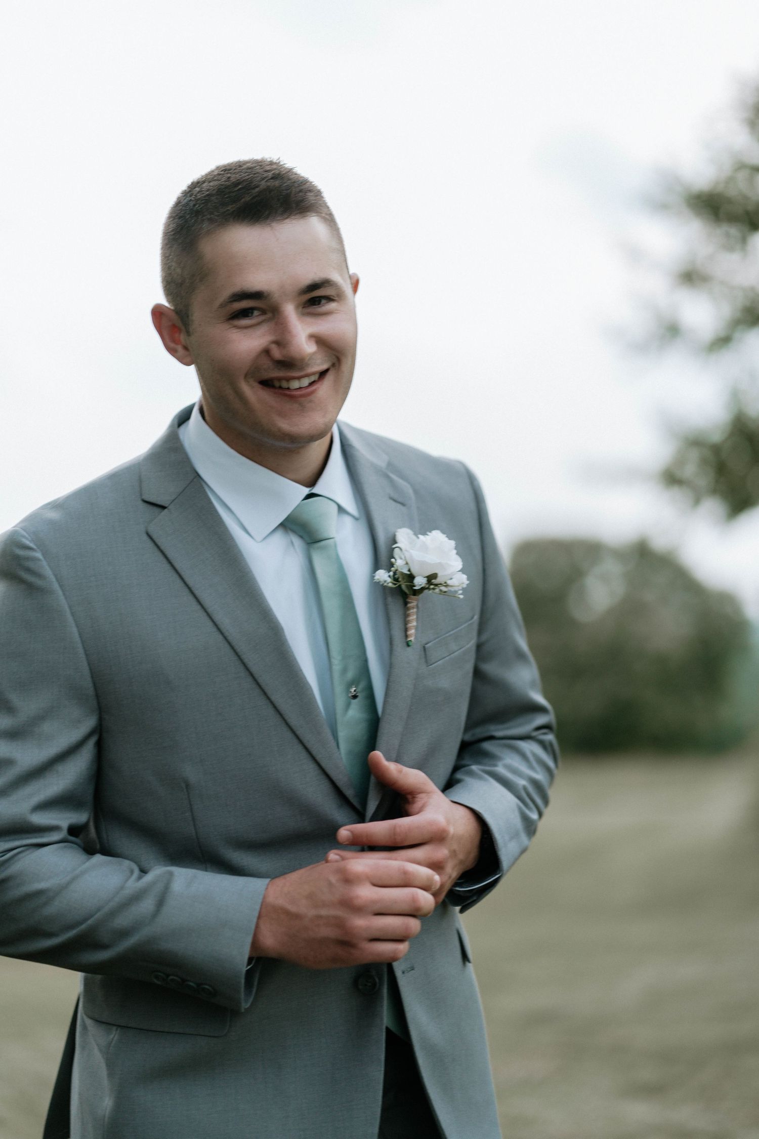 Smiling groom in gray suit with mint tie and white boutonniere posing outdoors at wedding.