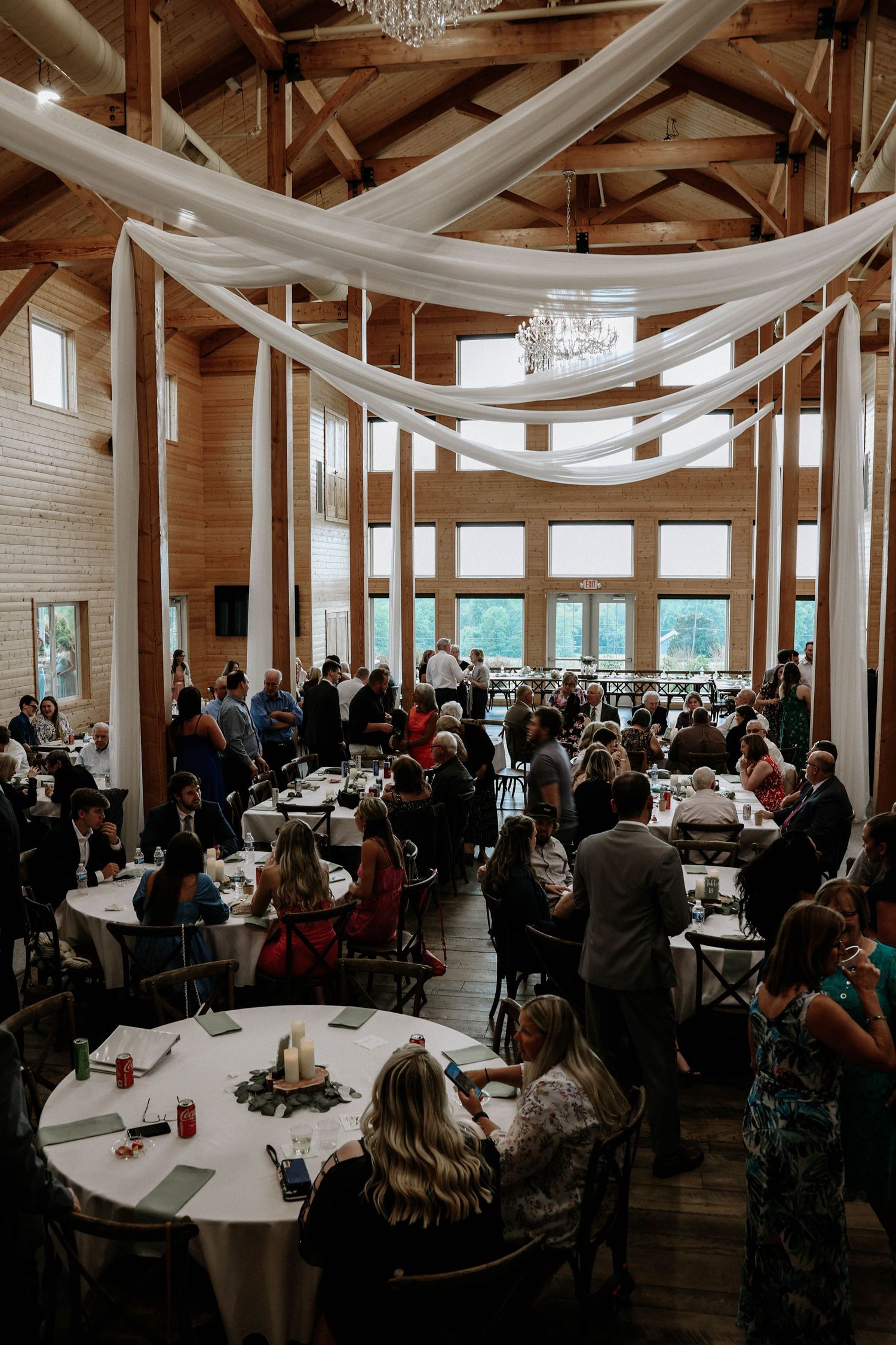 Rustic wedding reception hall with vaulted wood ceiling, white fabric draping, and chandelier filled with guests.