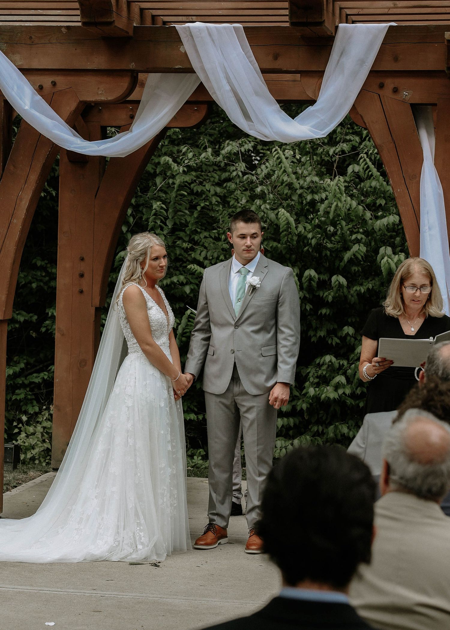 Bride and groom hold hands at outdoor wedding ceremony under wooden arch with white draping and lush greenery backdrop.