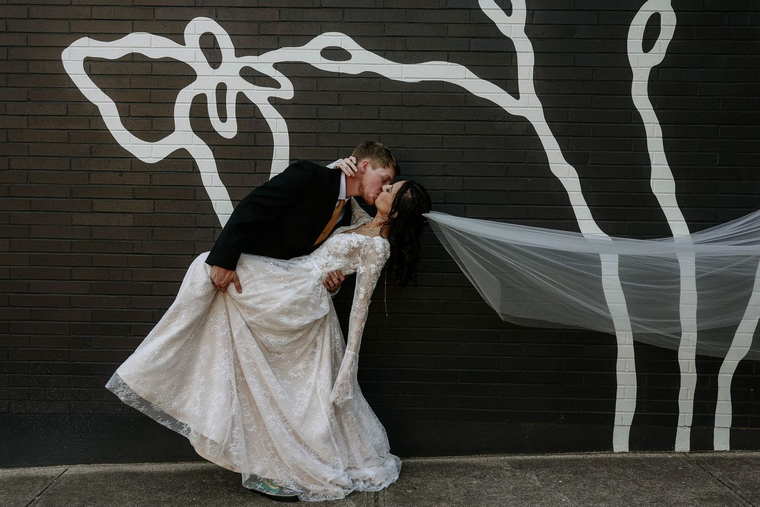 Bride and groom share a romantic dip kiss in front of a black brick wall with white cow mural art.