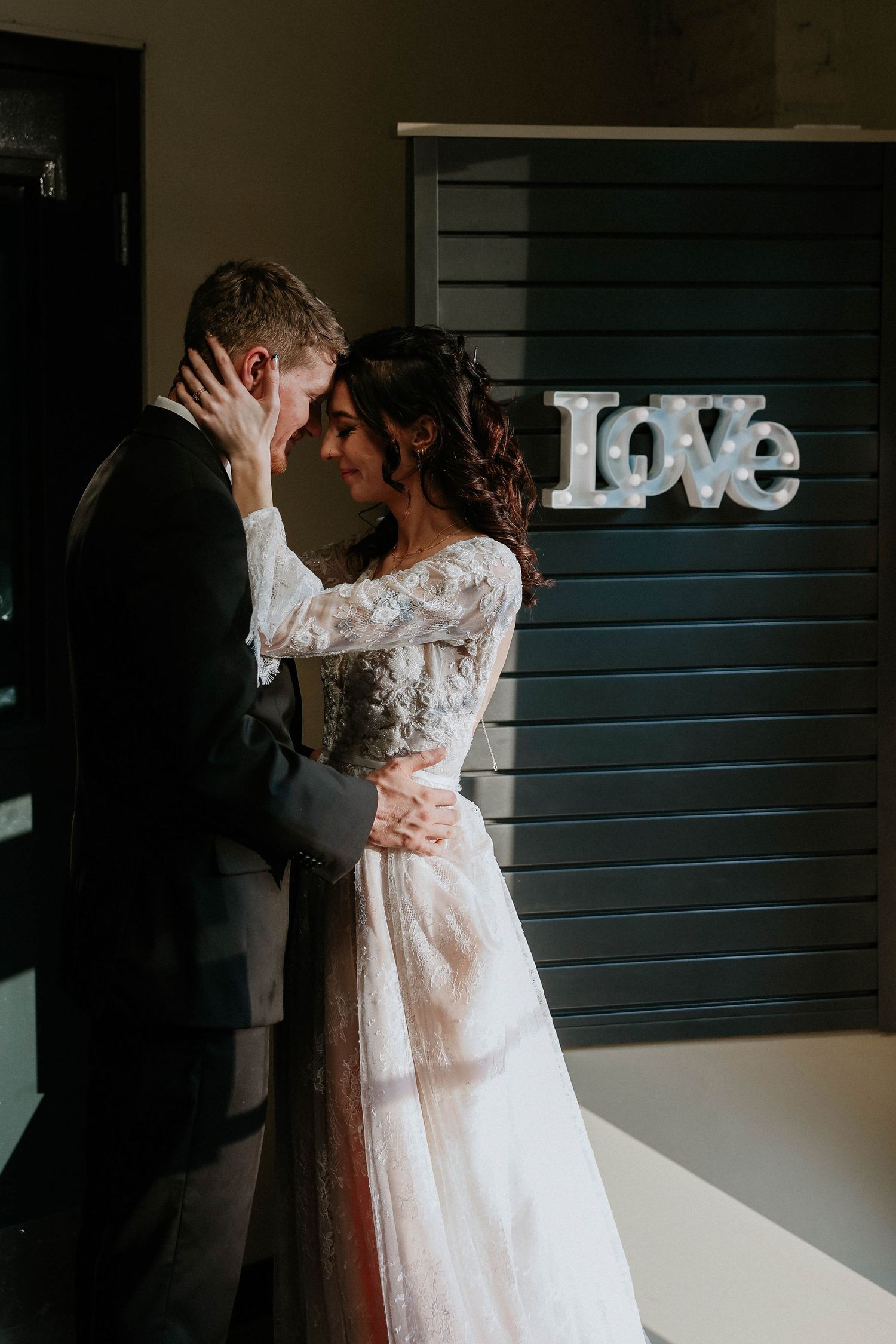 A bride and groom share a romantic moment in front of a glowing LOVE sign on their wedding day.
