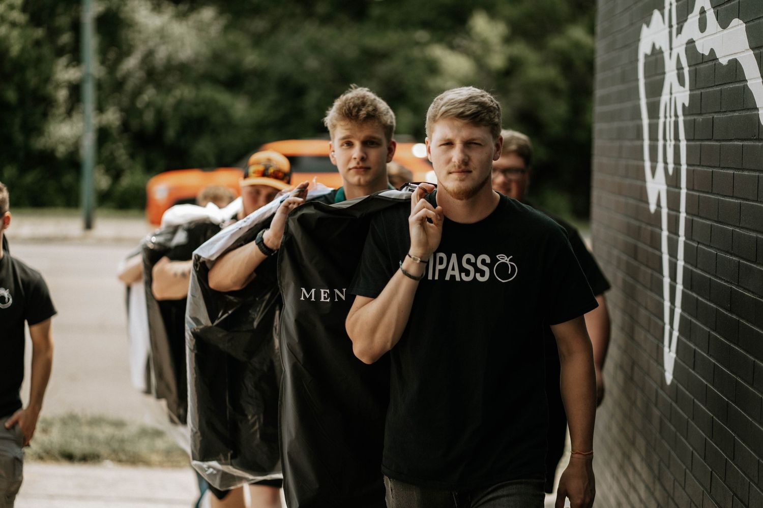 Two young men carrying black garment bags smiling while walking outdoors near a brick wall.