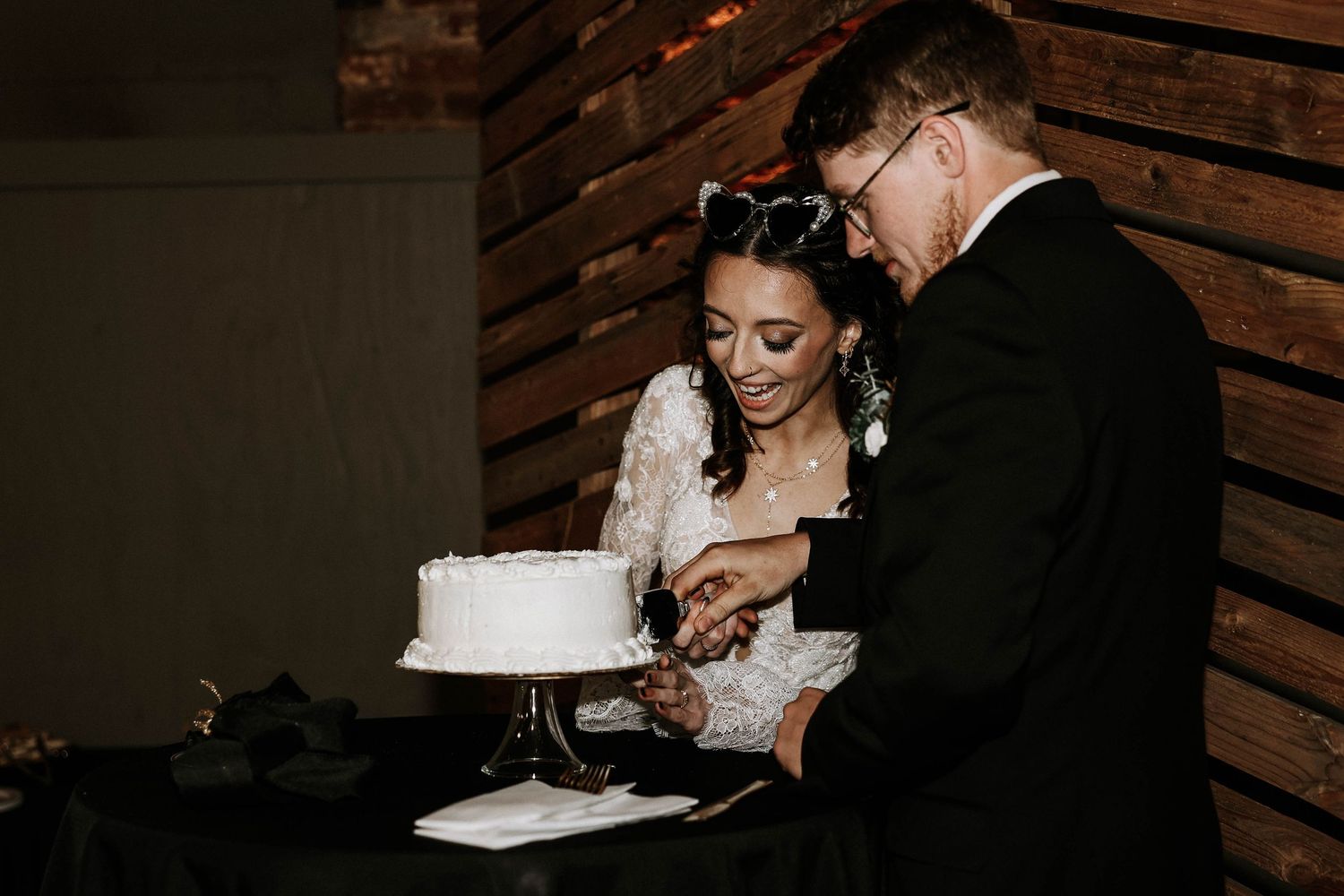 Bride and groom cutting their white wedding cake together at a romantic dimly lit reception venue.