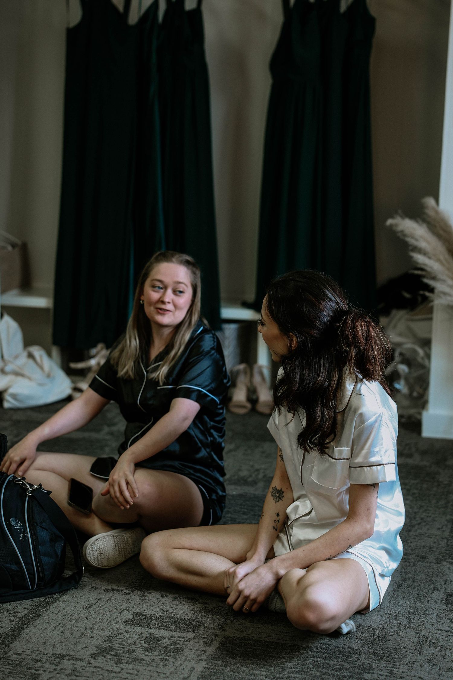 Two bridesmaids in satin pajamas sit on the floor chatting in a wedding preparation room with dark green dresses hanging behind them.