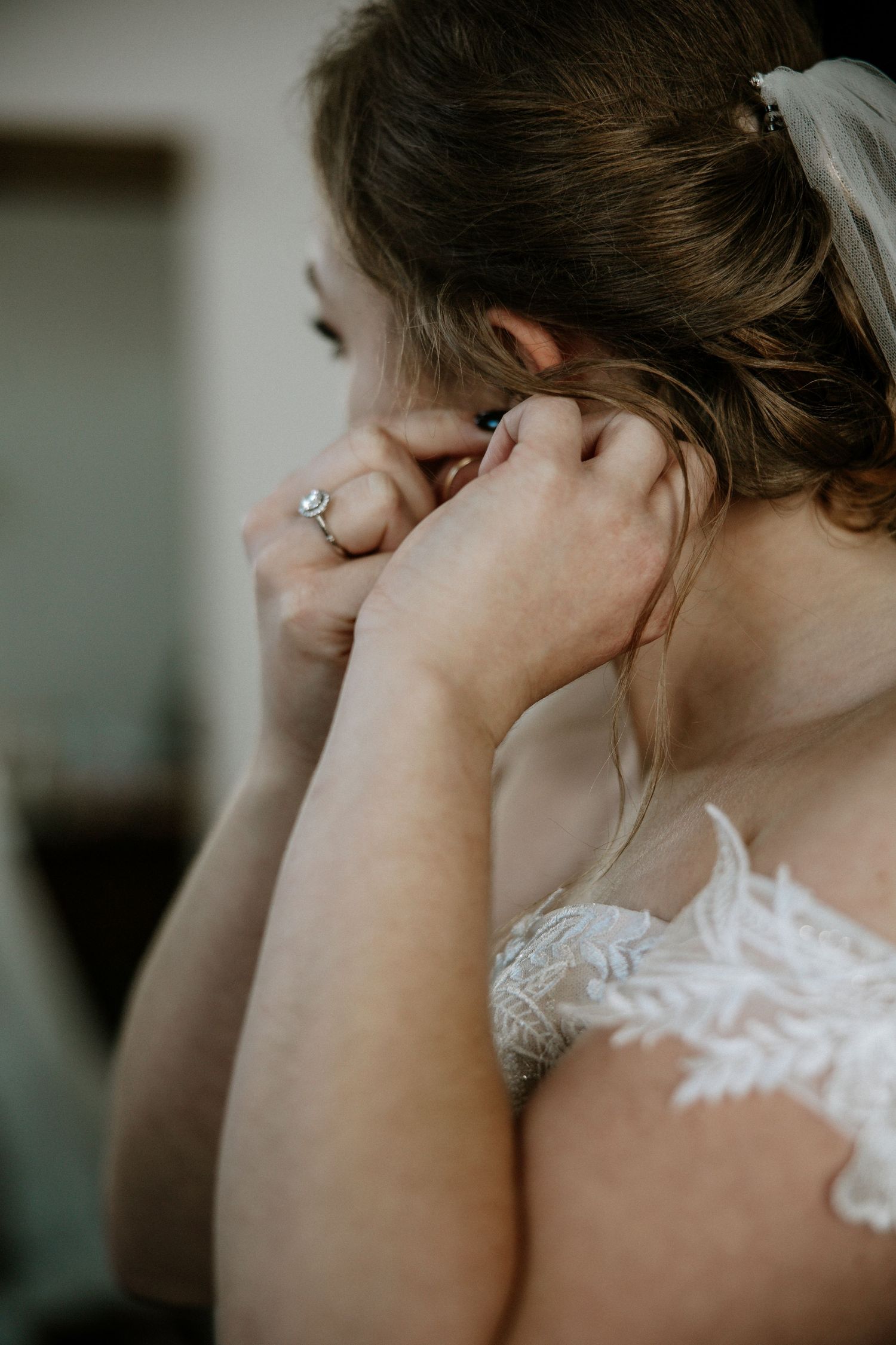 Bride putting on earrings while wearing an intricate lace off-shoulder wedding gown and veil at Urban Artifact in Cincinnati, Ohio