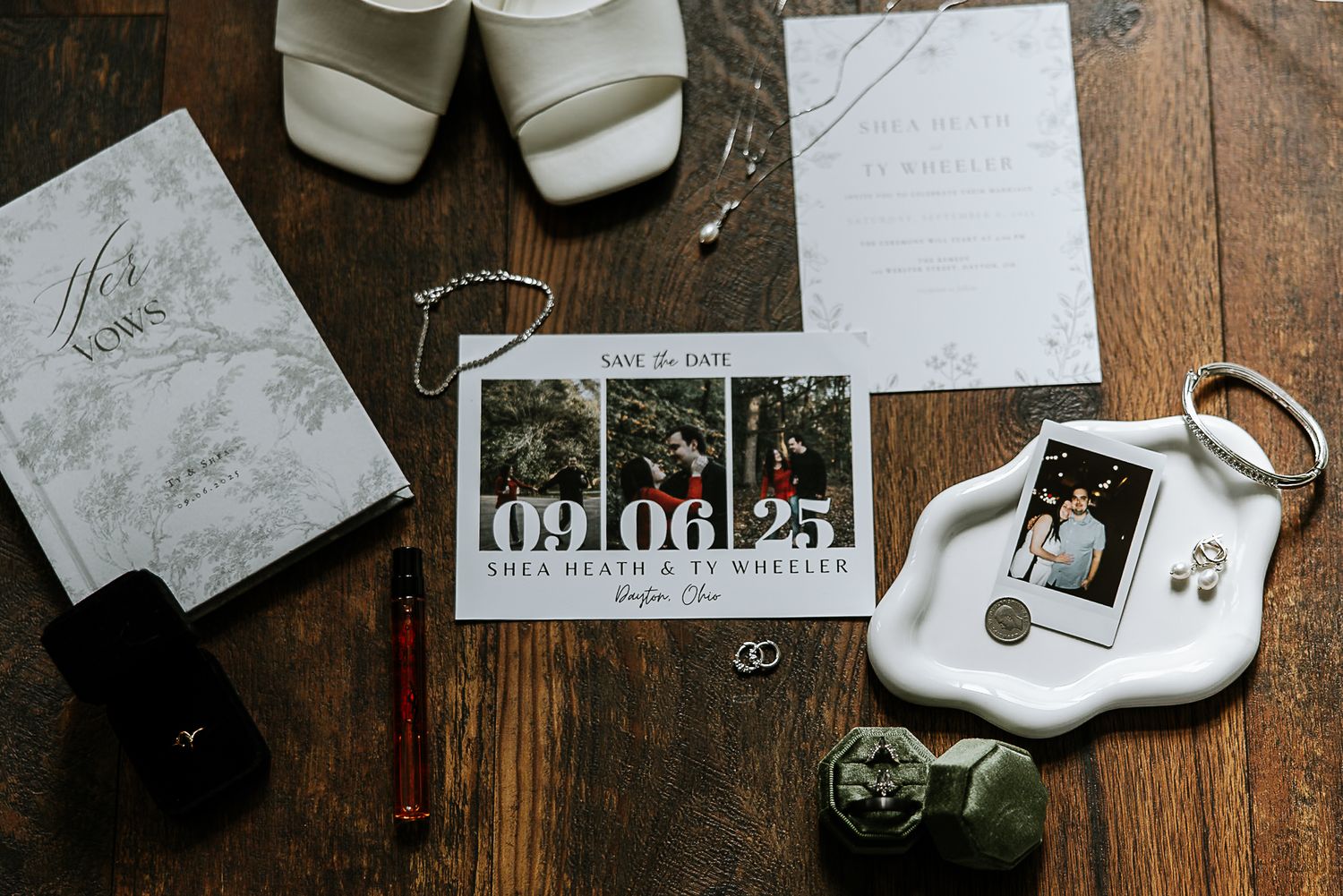 Wedding flat lay featuring save the date cards, white heels, rings, and bridal accessories on wood.