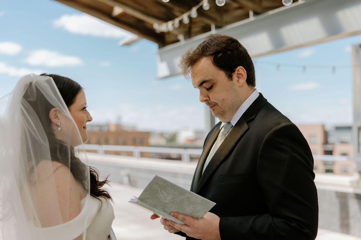 Groom reads handwritten vows to bride on sunny rooftop, both dressed in elegant wedding attire.