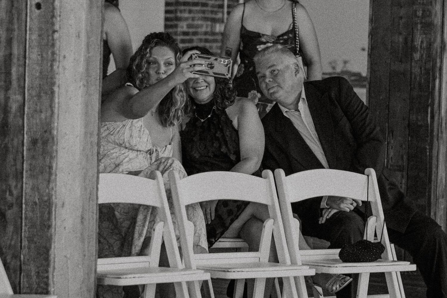 Three wedding guests taking a selfie together in black and white photo at ceremony.
