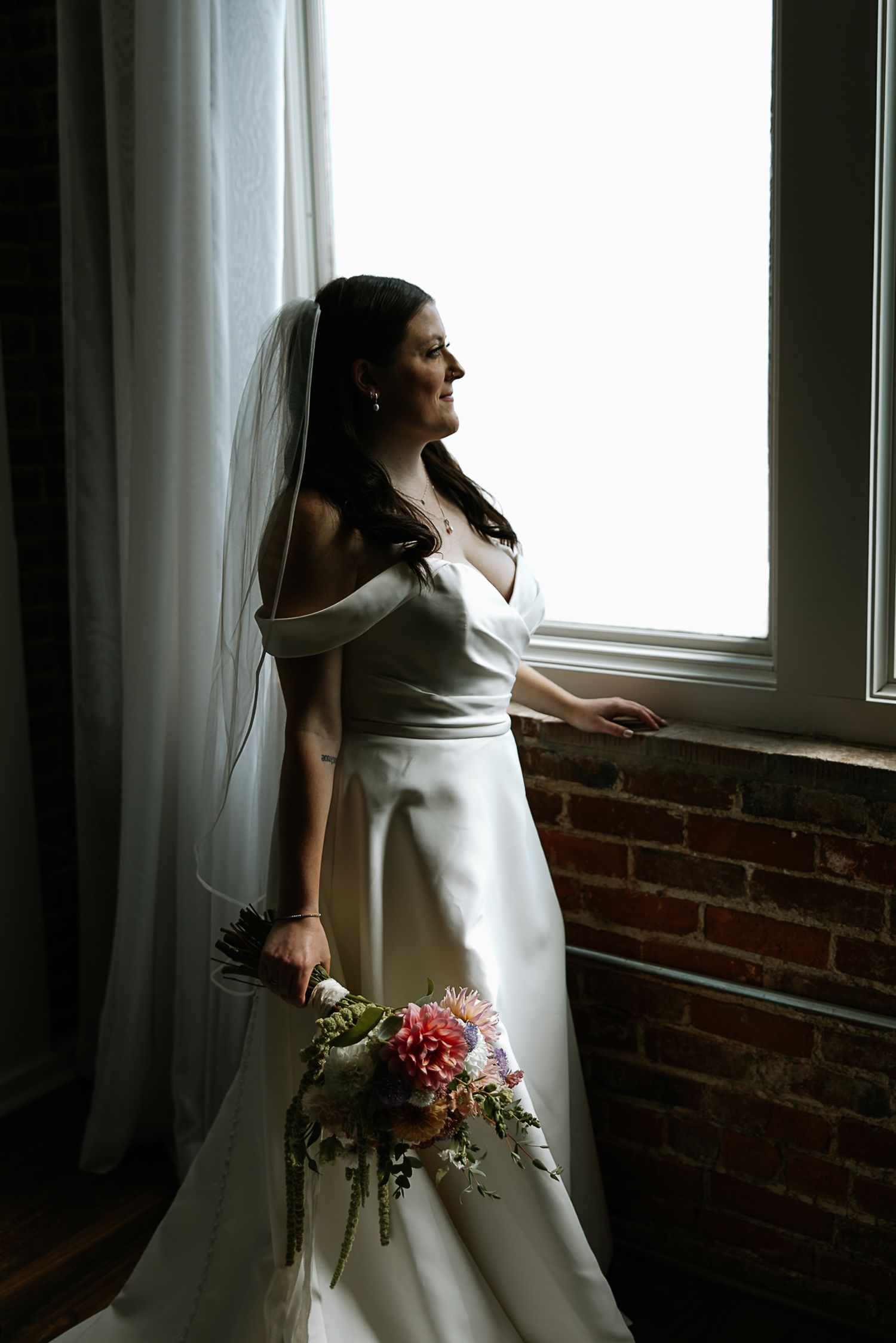 Bride in white off-shoulder gown and veil holding colorful bouquet, gazing out window with brick wall backdrop at The Remedy in Dayton, Ohio