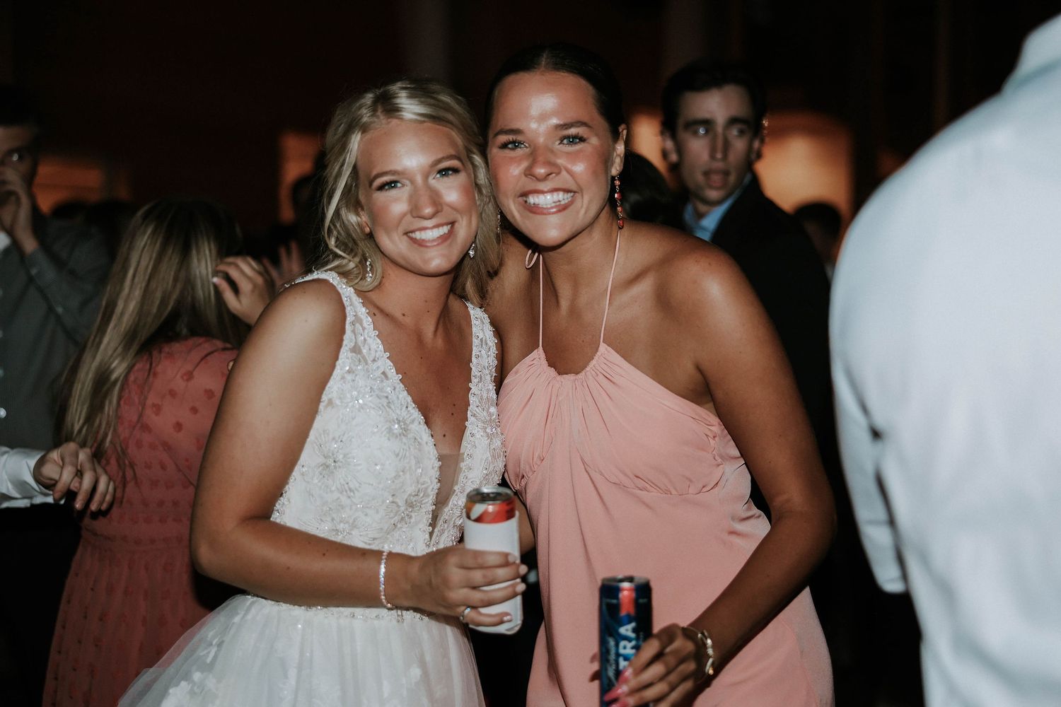 Two women smiling at a wedding reception, one in a white bridal gown and one in a pink bridesmaid dress, holding drinks.