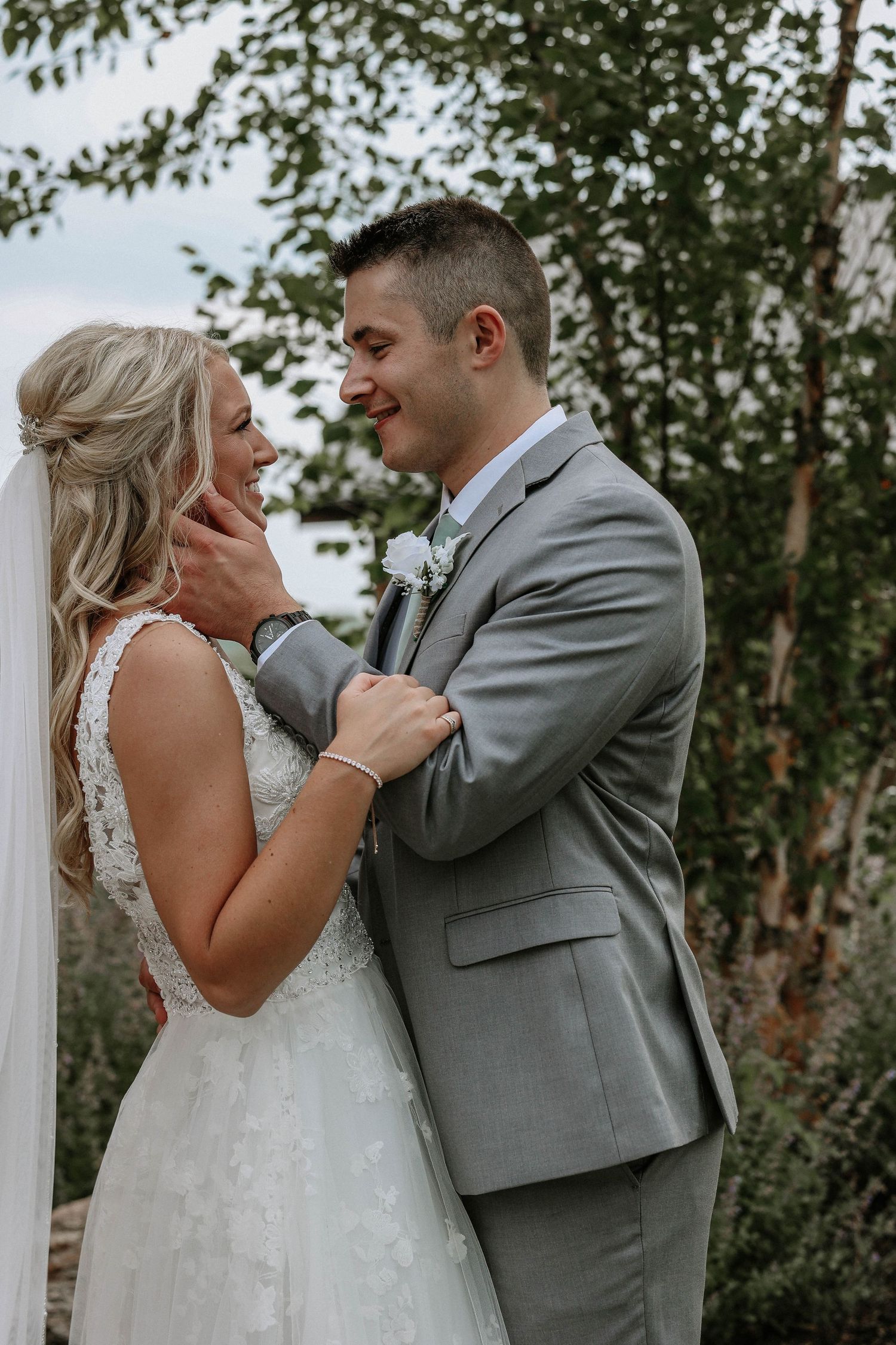 Smiling bride and groom share a tender moment outdoors, with the groom in a gray suit and bride in lace gown.