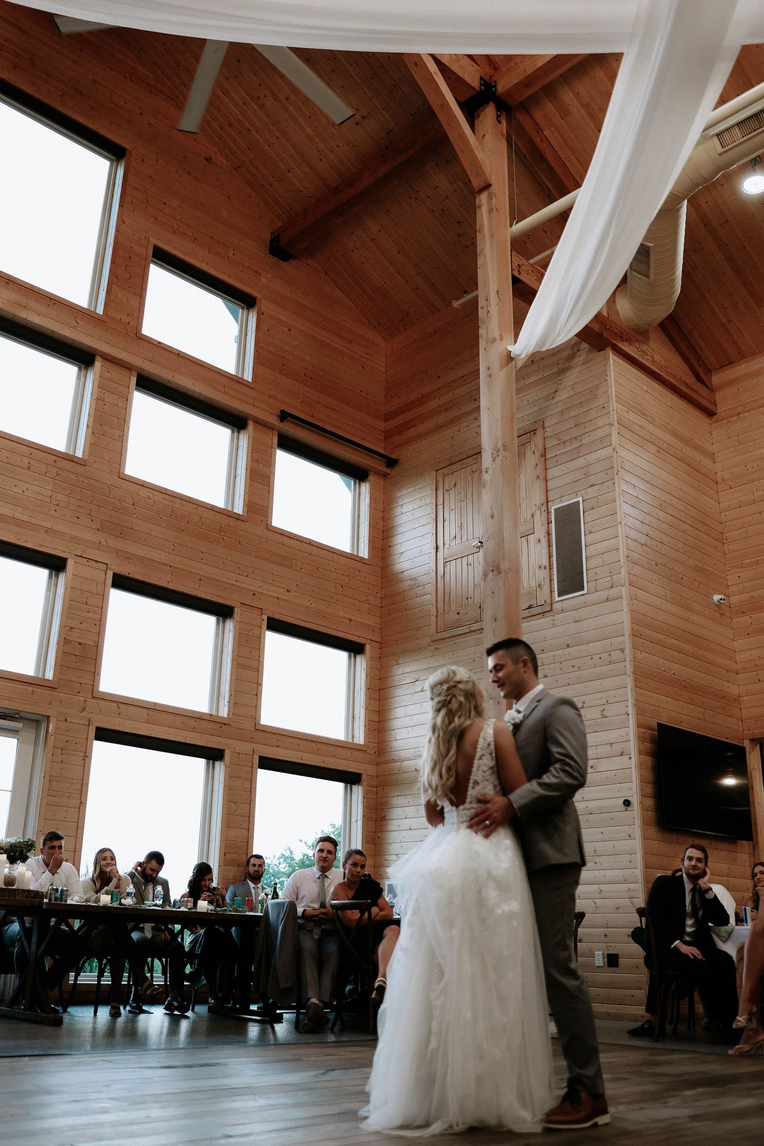 Bride and groom share first dance in rustic wood-paneled wedding venue with soaring ceilings and large windows.