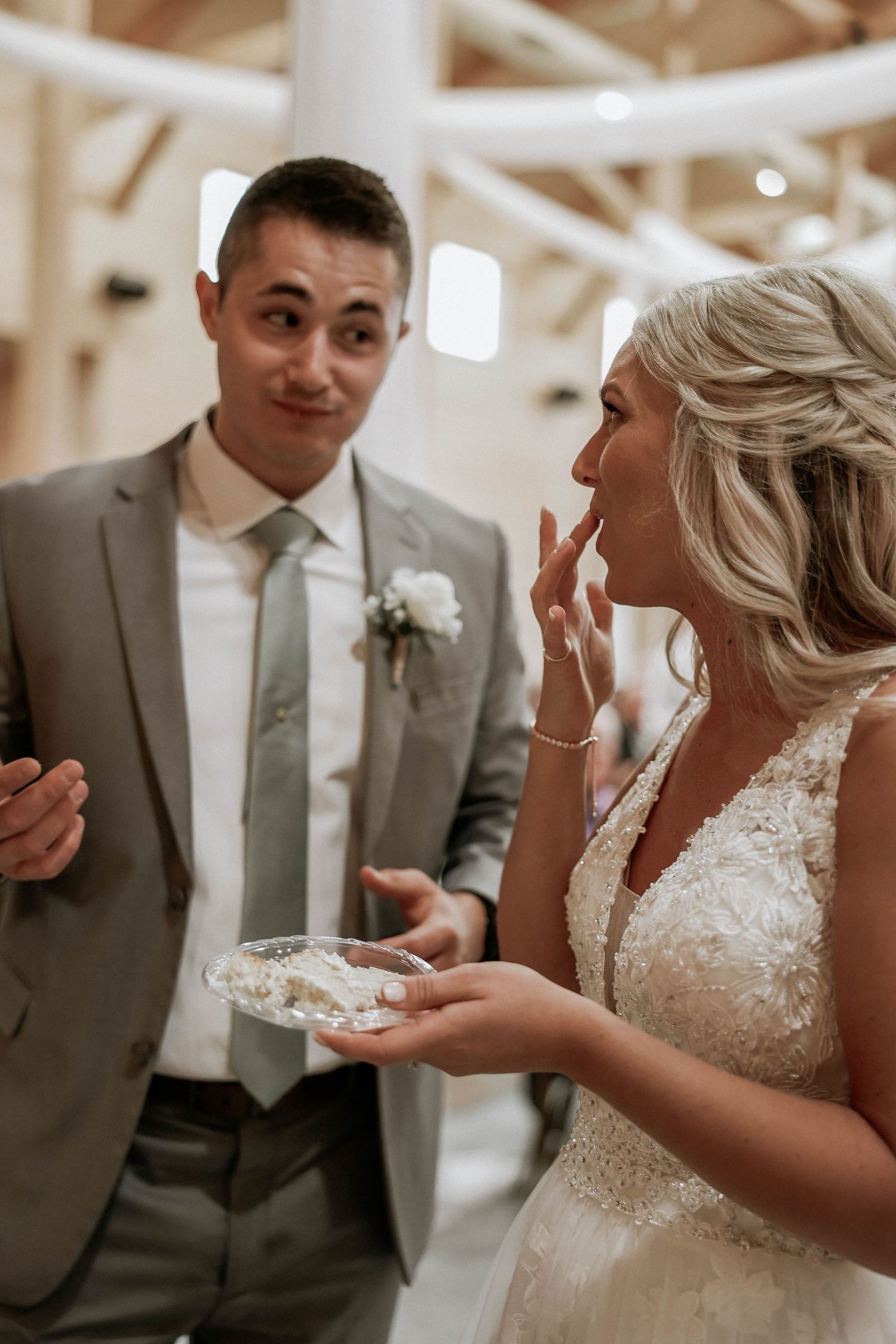 Bride and groom sharing wedding cake, smiling together in elegant barn venue, bride in lace gown, groom in gray suit.