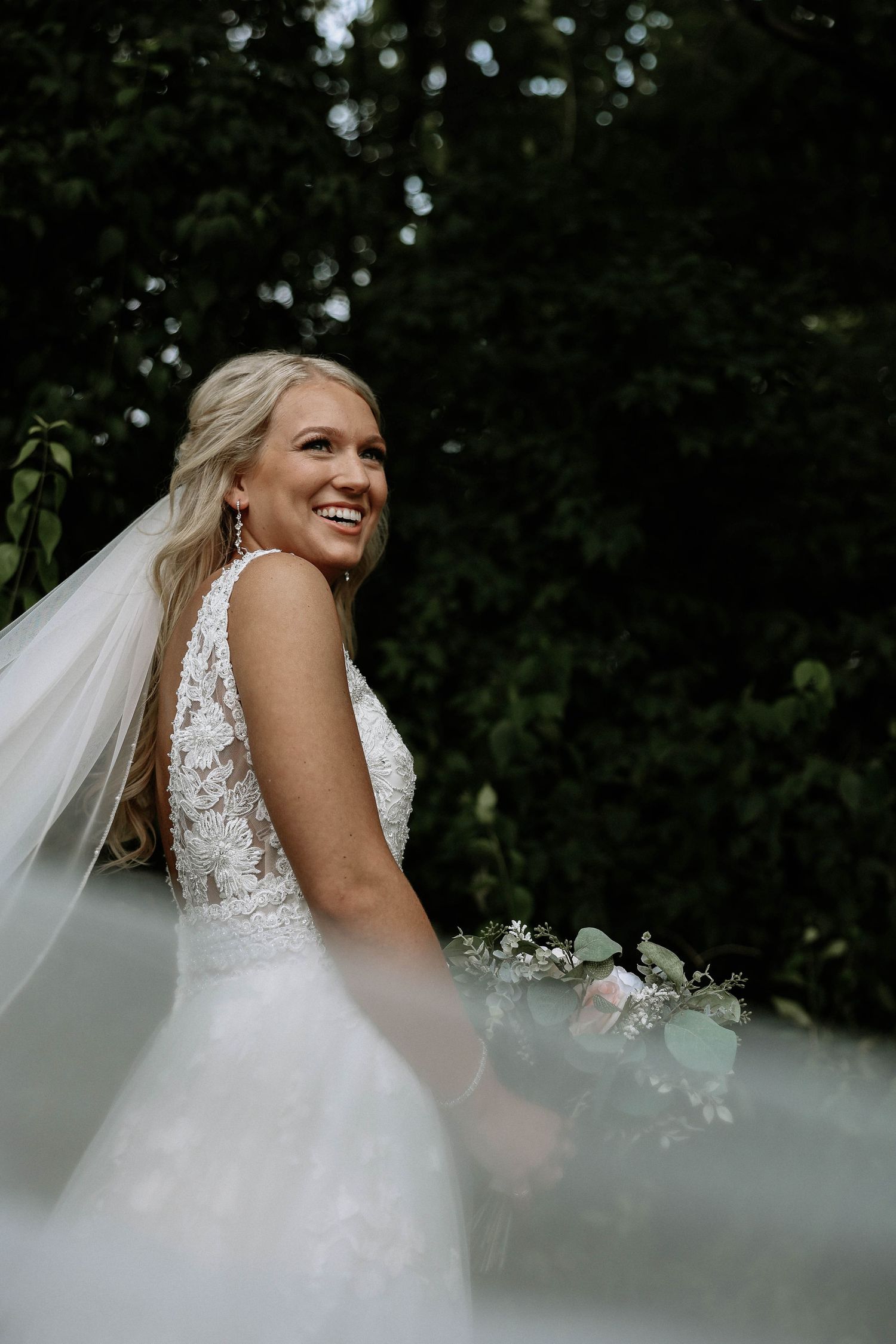 Radiant blonde bride in lace wedding gown smiling with veil flowing in breeze.