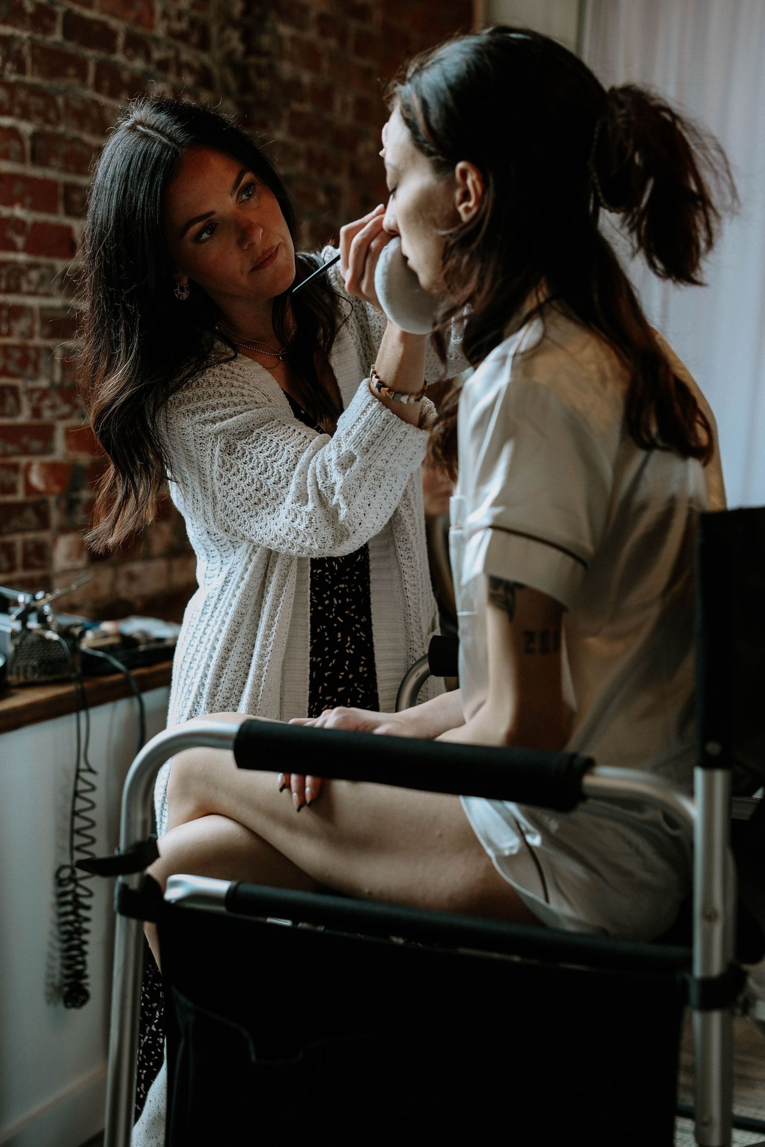 Makeup artist applying makeup to a bride seated in a chair in a rustic brick-walled room.