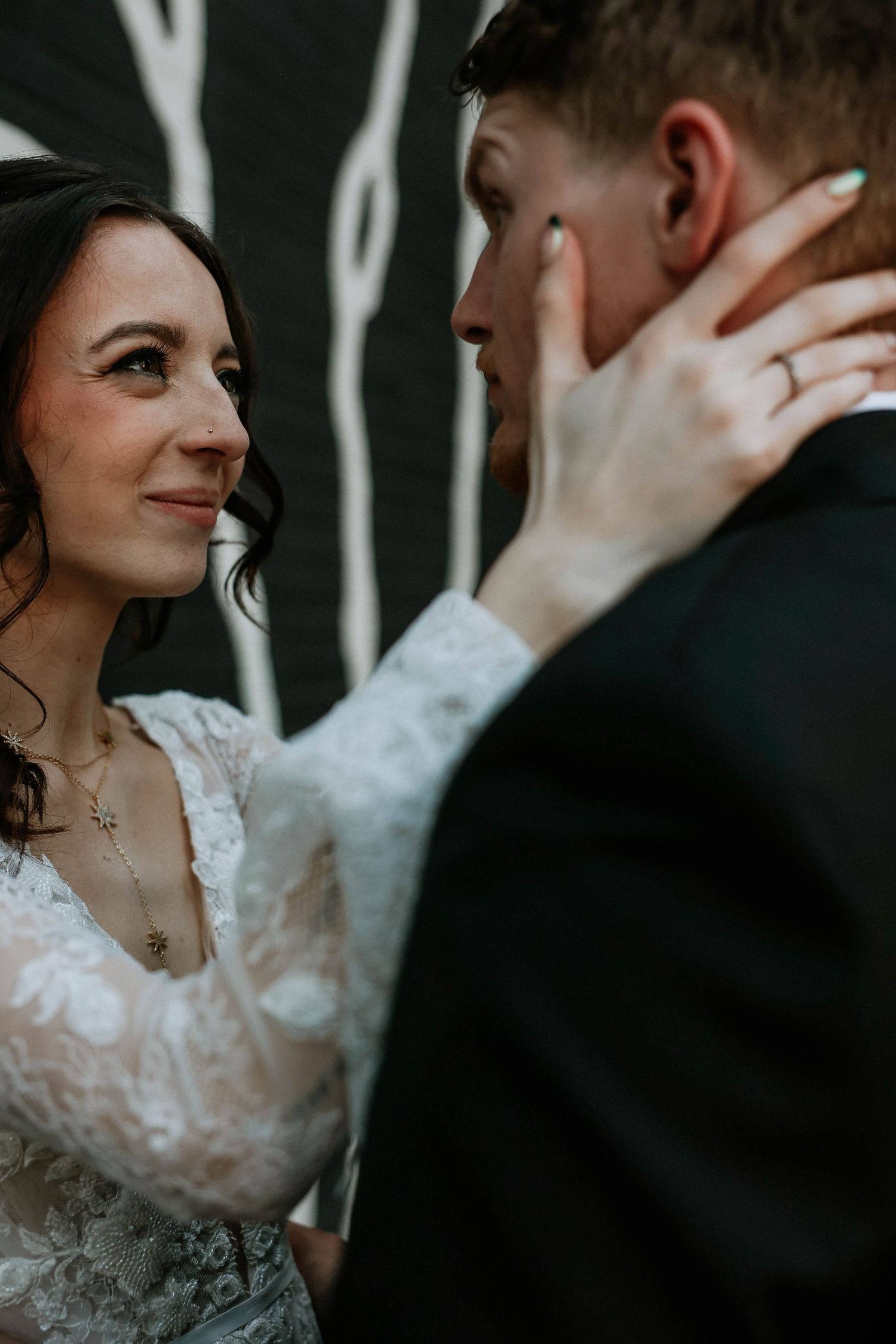 Bride in lace dress tenderly holds groom's face, sharing an intimate moment at their wedding ceremony.