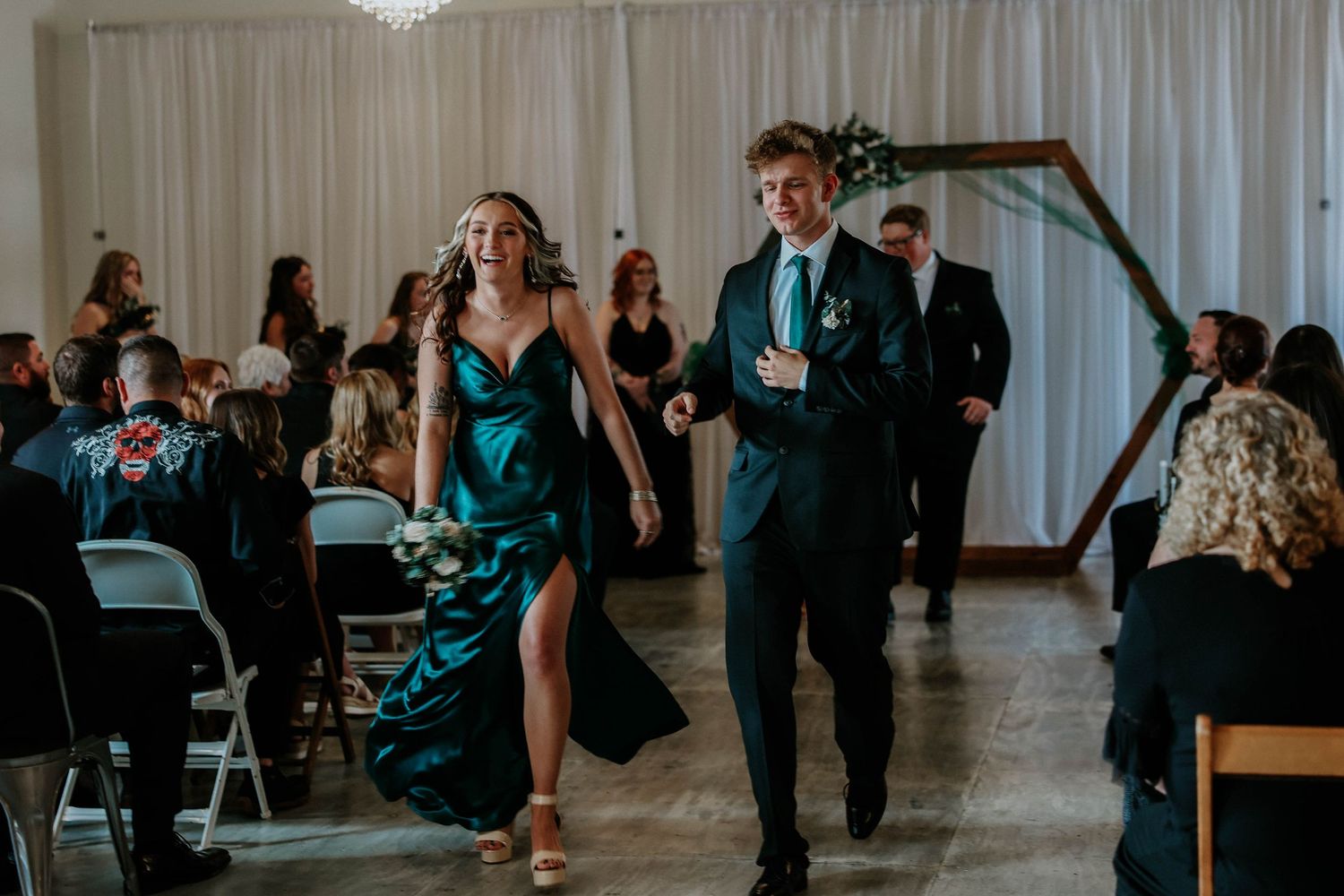 A bridesmaid in a teal gown walks down the aisle with a groomsman in a dark suit at an elegant indoor wedding.