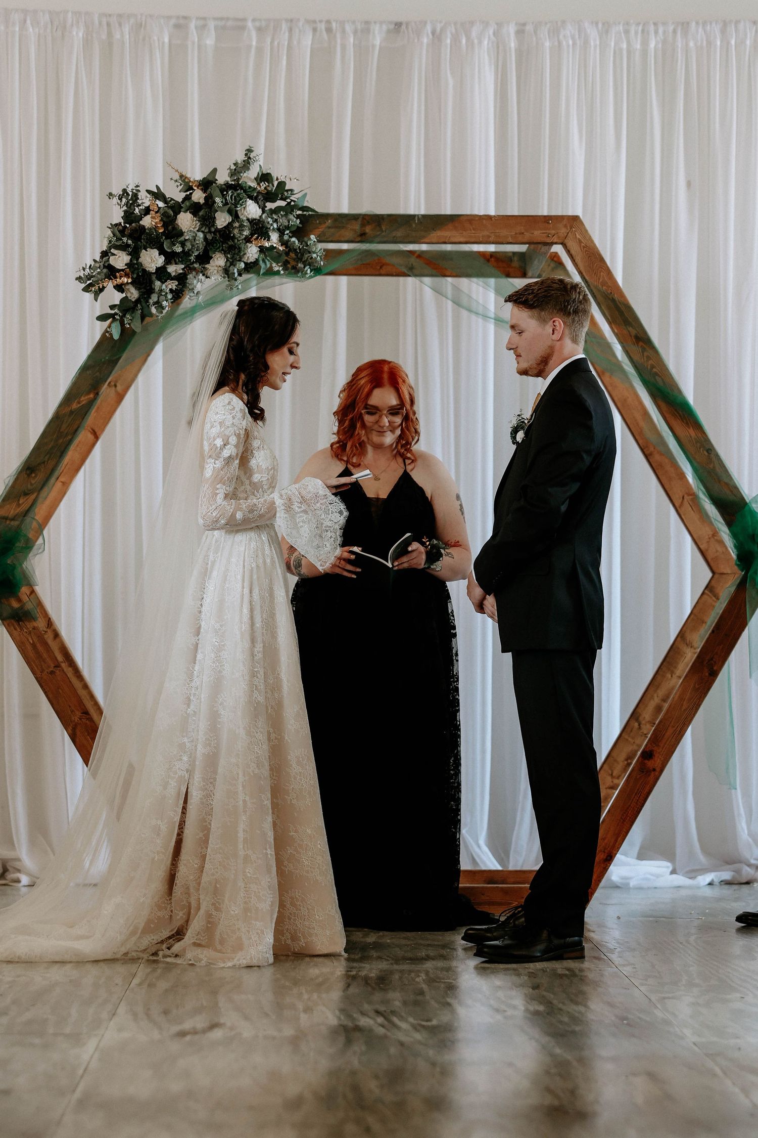 Bride and groom exchange vows before a wooden hexagon arch adorned with flowers, officiated by a red-haired woman in black.