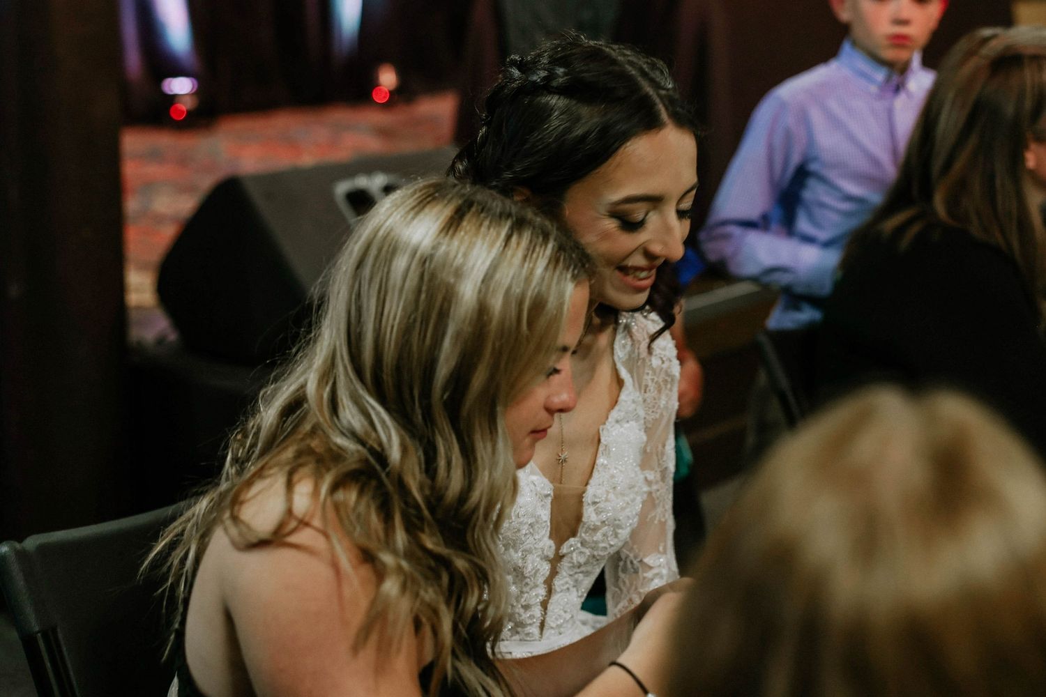 Bride in lace gown shares an emotional moment with a blonde guest at a wedding reception.