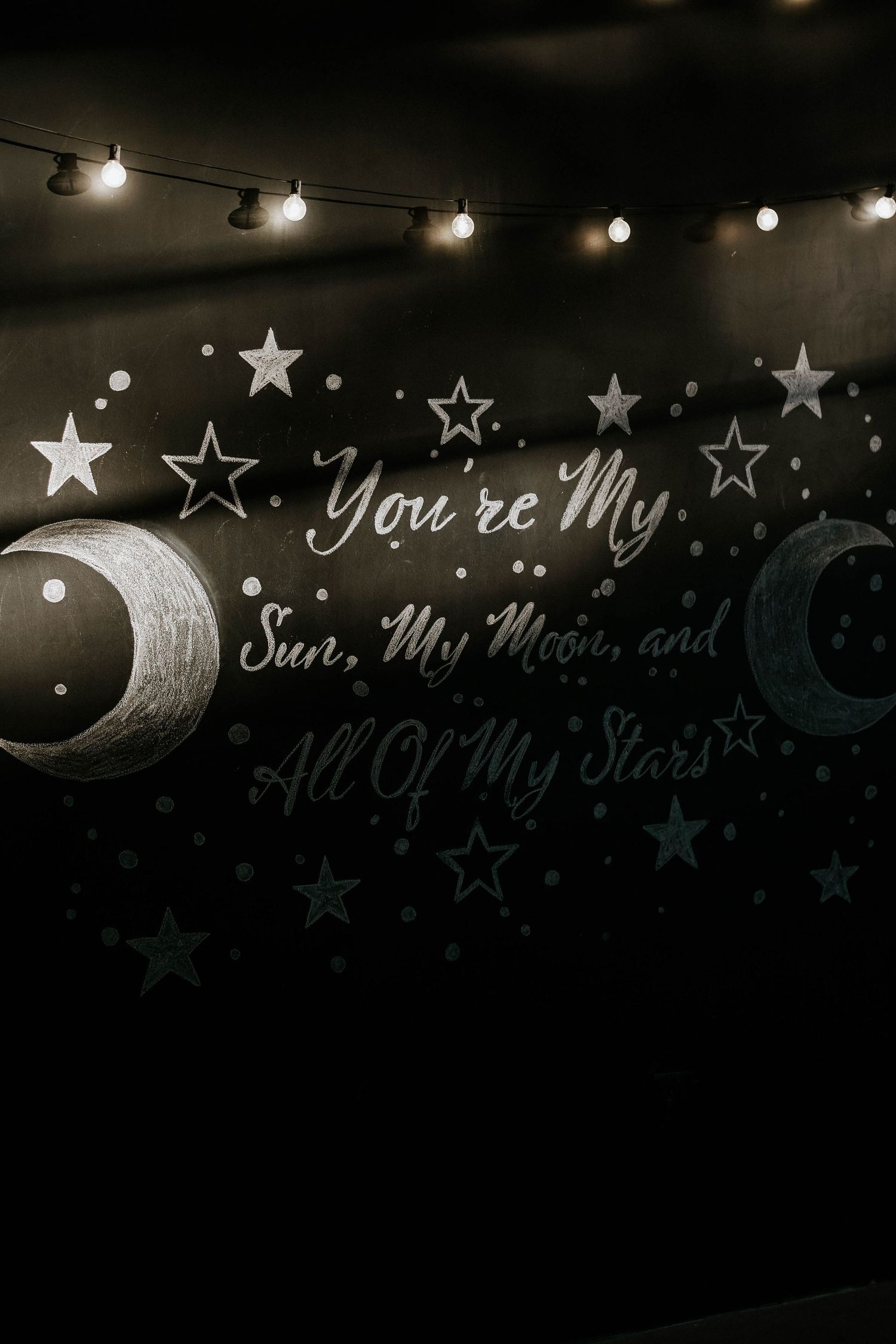 Dark wall mural with stars, moons, and the quote You're My Sun, My Moon, and All Of My Stars with string lights.