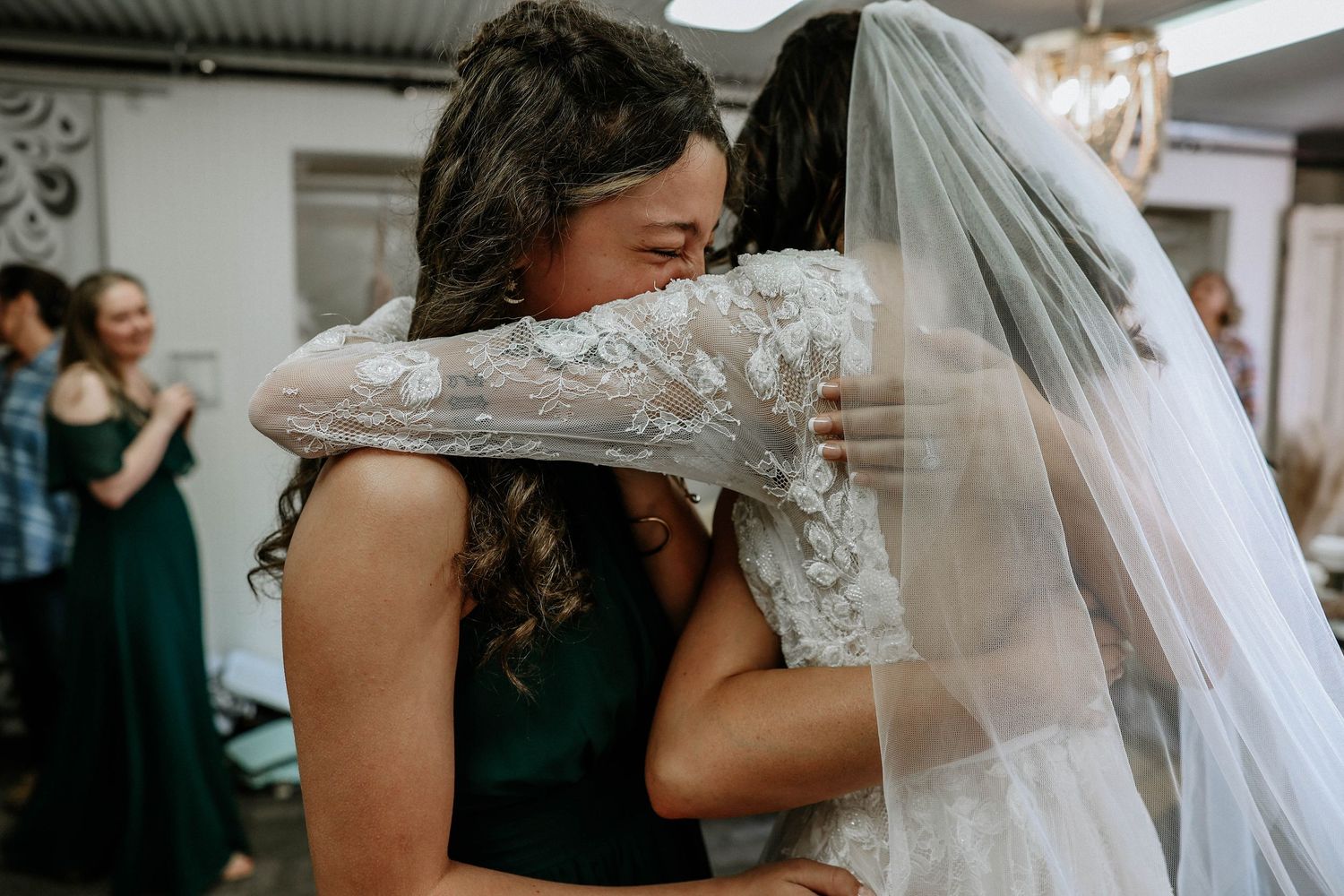 Bride in lace wedding dress and veil shares emotional hug with bridesmaid in green dress at wedding reception.