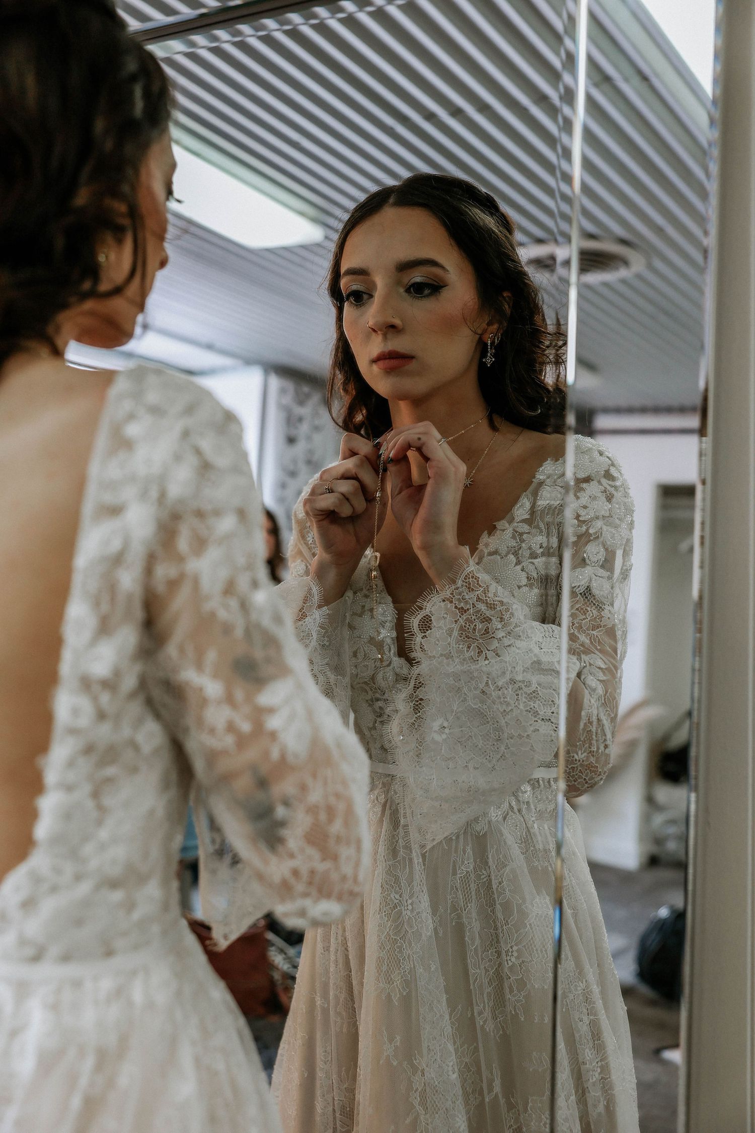 Bride in lace long-sleeve wedding dress adjusting earring while gazing into mirror during bridal preparation.
