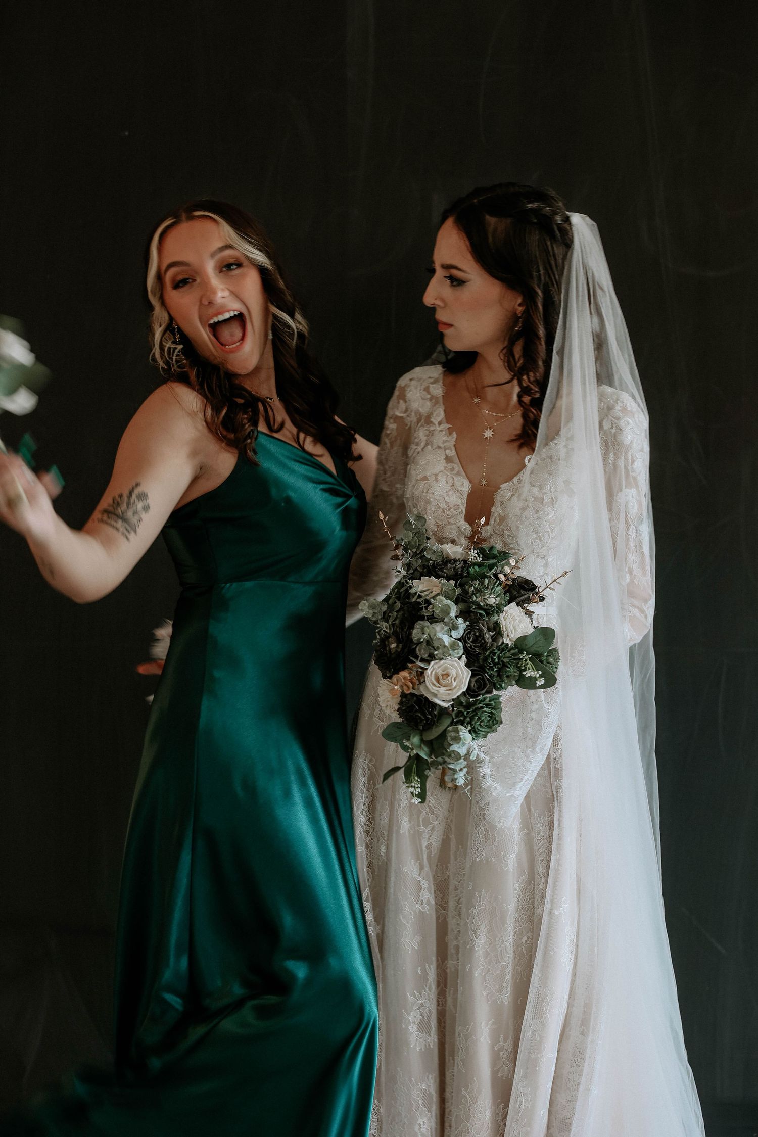 Bride in lace gown with veil holds bouquet beside bridesmaid in emerald satin dress celebrating joyfully.