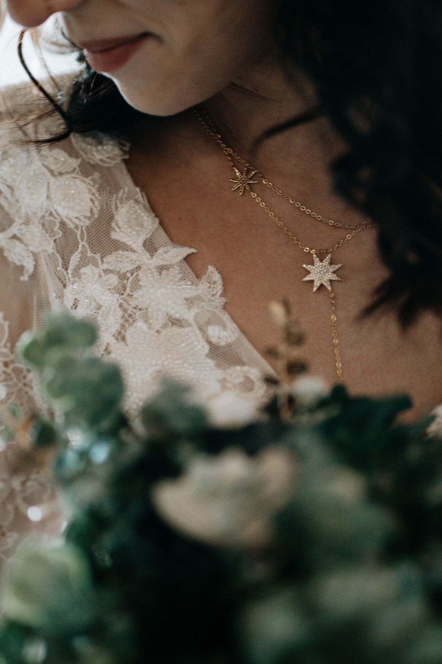 Bride wearing layered gold star necklaces over lace wedding dress, holding a green and white floral bouquet.