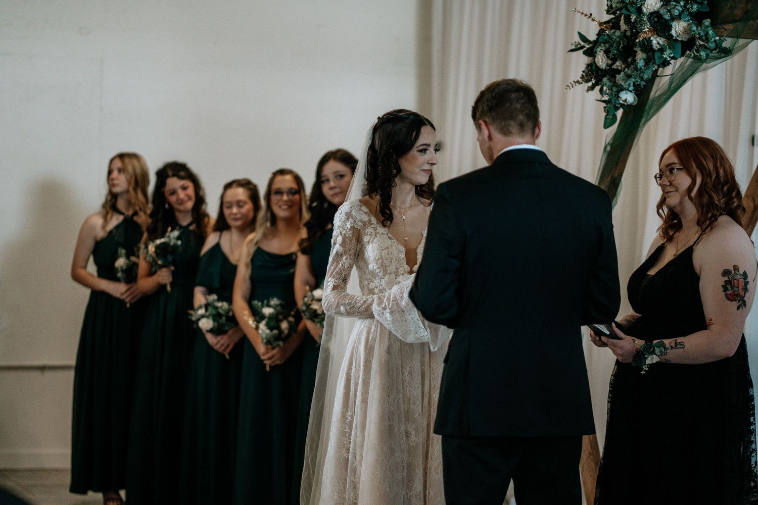 Bride and groom exchanging vows at altar surrounded by bridesmaids in dark green dresses at wedding.