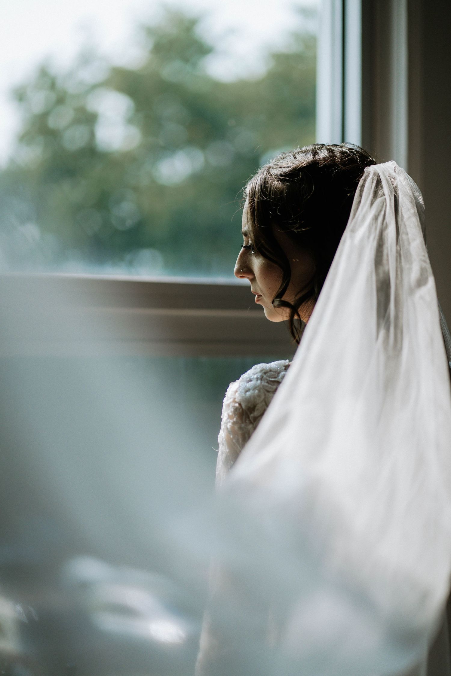 A bride in a white lace gown and veil gazes thoughtfully out a window on her wedding day at The Brightside in Dayton, Ohio