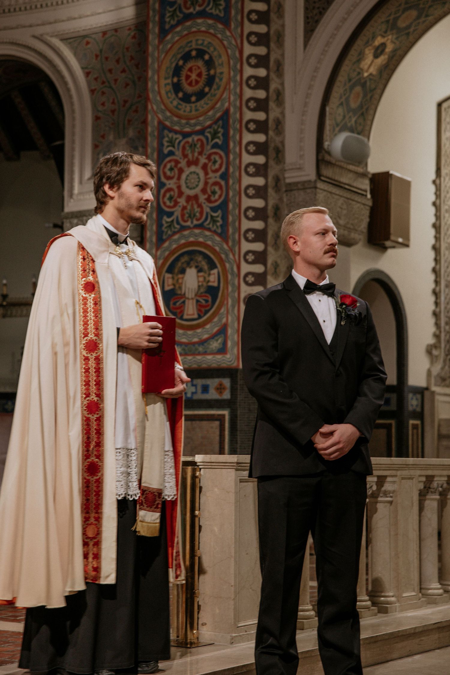 Priest in white ceremonial robes stands beside a groom in a black tuxedo at a church altar in Cincinnati, Ohio